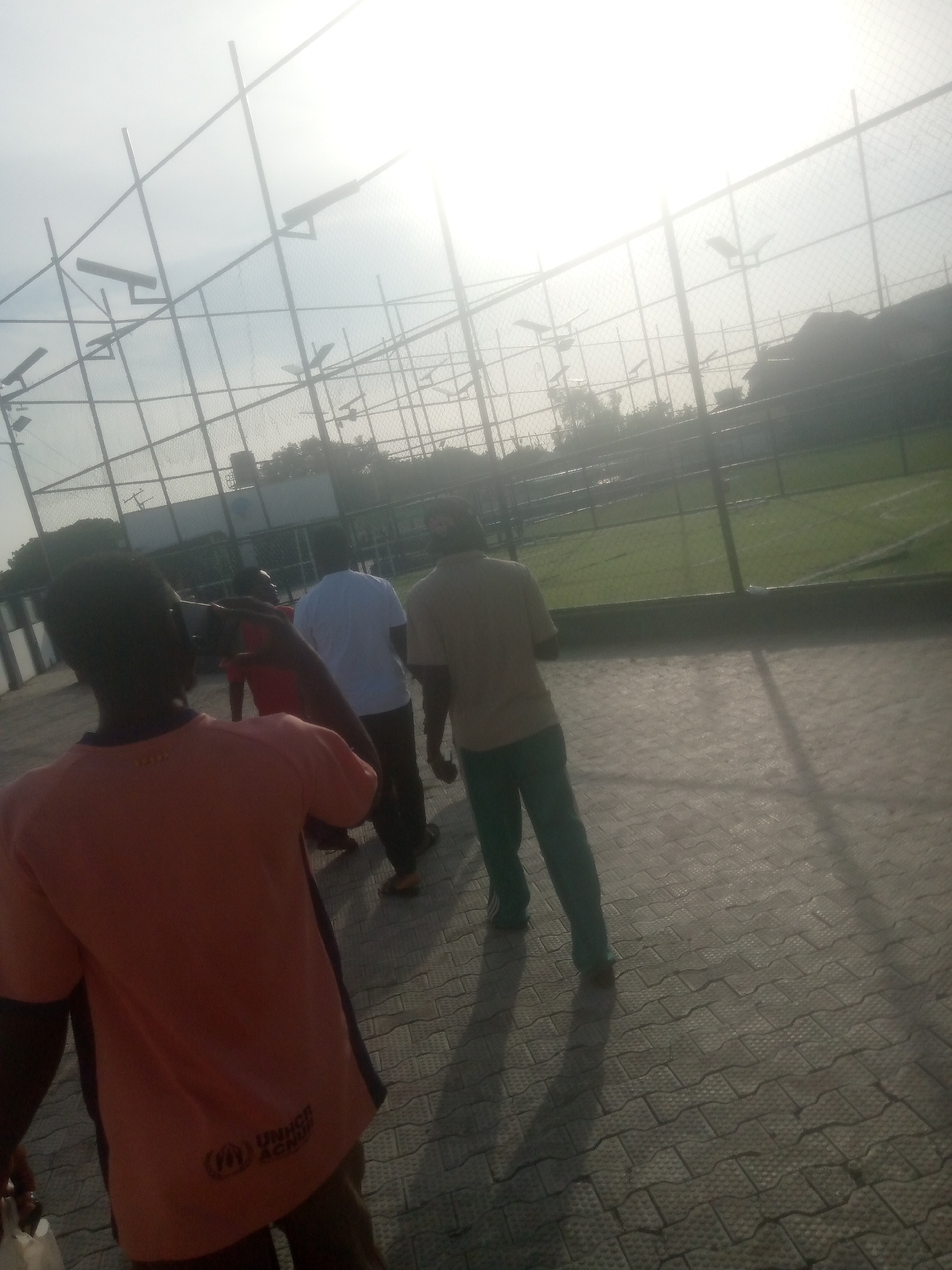 The image depicts a group of four individuals walking on a paved area adjacent to a fenced sports field. The field appears to be a soccer pitch with visible white markings. The fencing surrounding the field is constructed from metal poles and mesh. Overhead, a bright sky with a strong sun is visible, creating a glare and silhouette effects on some of the subjects and structures. Several floodlights are mounted on poles around the perimeter of the field.
In the foreground, a person wearing an orange t-shirt with a dark trim is walking away from the camera. The t-shirt has a UNHCR (United Nations High Commissioner for Refugees) logo on the back. To the right of this individual, another person in a light brown or tan t-shirt and green pants is walking in the same direction. Further back, a person in a white t-shirt and dark pants is visible, and behind them, a person in a red shirt. The paved ground is made of interlocking grey tiles. The overall impression is an outdoor setting, possibly a sports complex, in Jalingo, Nigeria, under bright sunlight.