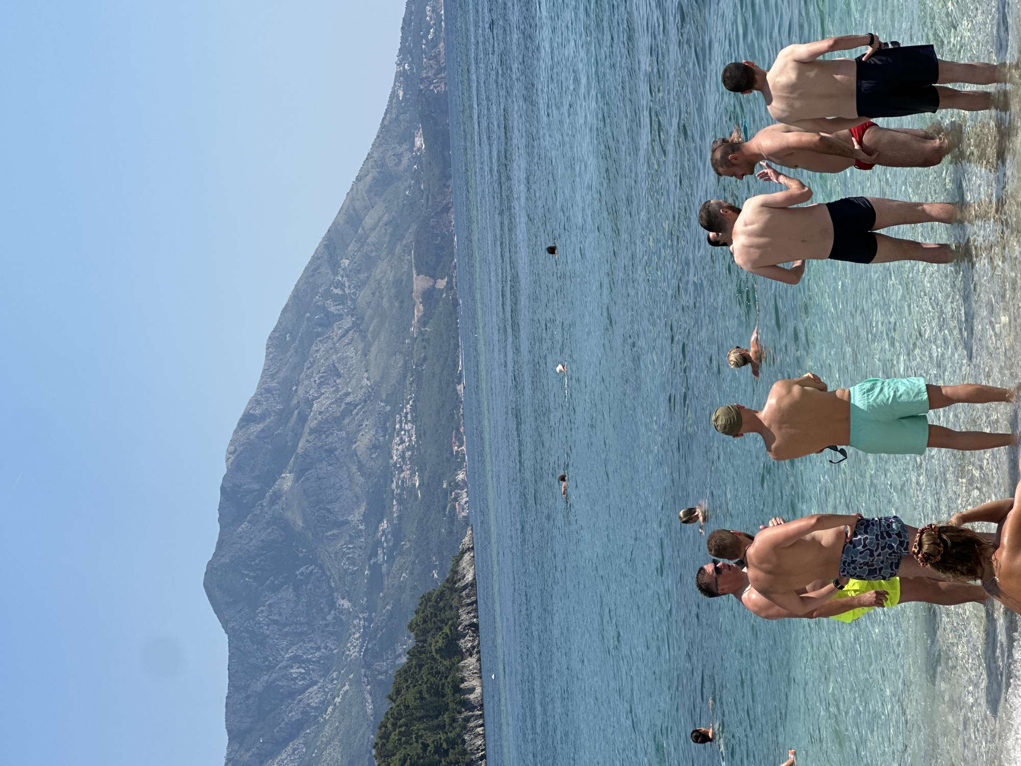 This media file shows a group of people on a sunny day at a beach in Budva, Montenegro. The scene is dominated by the clear blue water of the Adriatic Sea and a steep, mountainous landscape in the background, densely covered with green vegetation. Several individuals are partially submerged in the shallow, transparent water, appearing to be enjoying the sea and the warm weather.
The subjects are mostly men, dressed in swim trunks, and are engaged in casual activities, some standing in conversation, others seemingly walking through the water. One person is swimming further out in the sea. The overall mood appears relaxed and leisurely. The bright sunlight suggests it is midday. The clear sky and calm water indicate good weather conditions. The mountainous coastline with scattered buildings hints at the urban proximity of the beach. There is no visible text in the image.