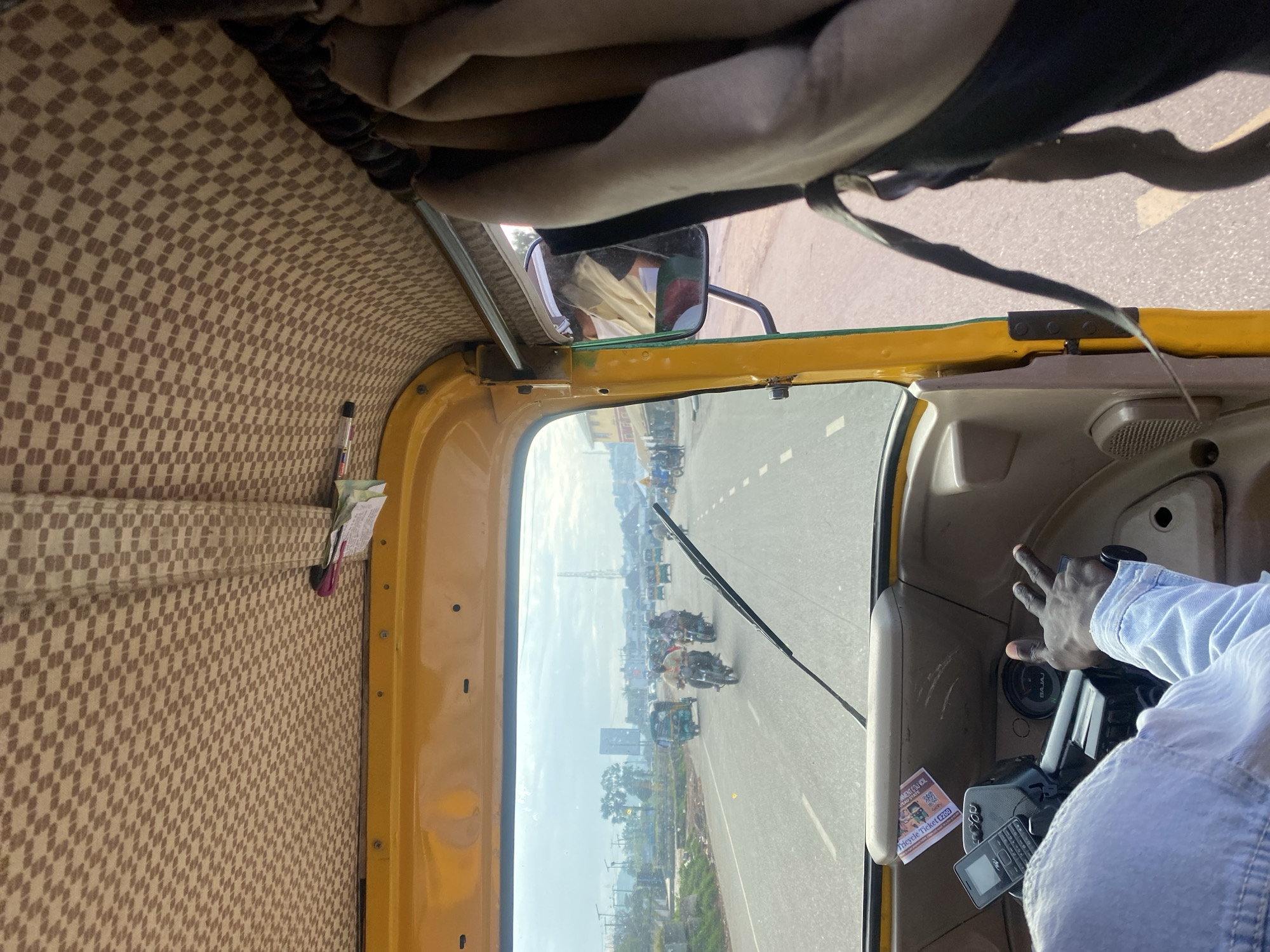 The image is taken from the interior of a yellow auto-rickshaw, looking forward through the windshield. The ceiling of the auto-rickshaw is covered with a beige and brown checkered fabric. A beige curtain is visible on the right side, hanging down. The driver's hands are visible in the lower right of the frame, with one hand on the steering wheel and the other raised in a peace sign. A mobile phone is mounted on the dashboard. Outside, a multi-lane road is visible with several other auto-rickshaws, motorcycles, and some cars. Buildings and infrastructure, including a tall tower, are seen in the distance. The environment appears to be urban or suburban, with clear skies. The location is identified as Rafin Par, Nigeria.
