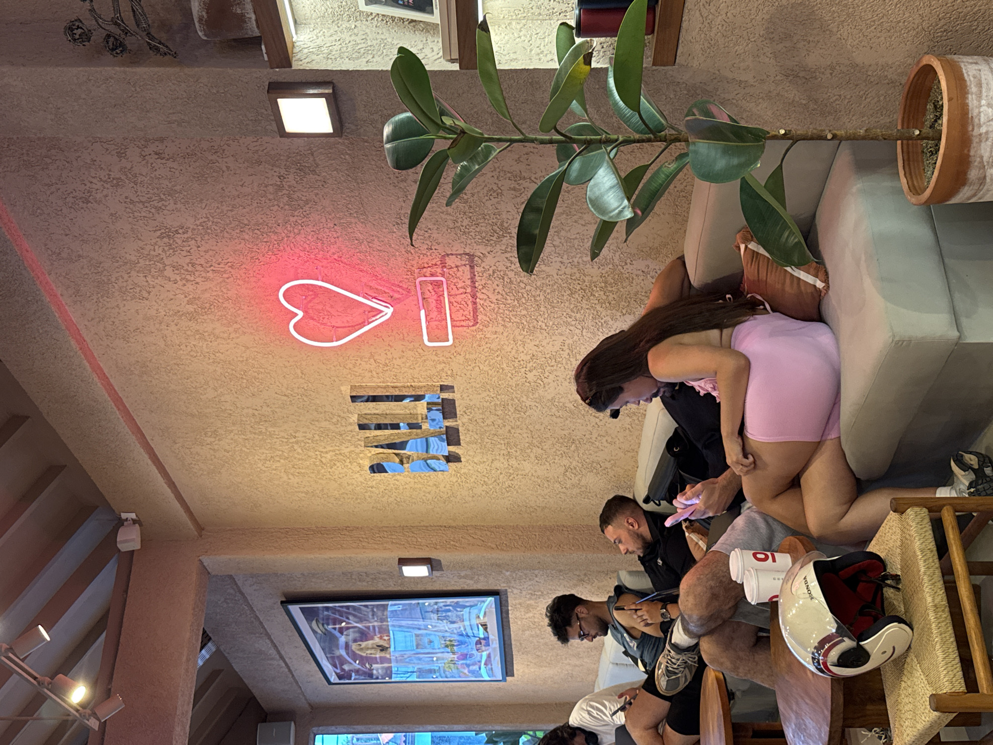 The image captures an indoor scene, likely a cafe or lounge, featuring several young adults engaged in casual activities. The setting includes textured walls and ceiling, with prominent structural beams and warm artificial lighting from square wall-mounted fixtures and track lights.
In the foreground, a woman wearing a pink top and shorts is seated on a light-colored sofa, looking down at her mobile phone. Her long, dark hair cascades down her back. Next to her, a man in a black shirt is also engrossed in his phone. Other individuals, primarily men, are visible further into the frame, some also using phones, while one appears to be writing or drawing.
Notable objects include a large potted plant (Ficus elastica) with broad, dark green leaves, a distinctive pink neon sign on the wall depicting a heart shape and a vertical bar (possibly part of "LOVE" or "LI"), and a framed piece of artwork on the wall. A white and red motorcycle helmet, clearly displaying "HONDA" on its side, rests on a wooden chair next to two disposable coffee cups that show a partial "LO" or "10" logo. A brown handbag is placed on the sofa.
The people appear relaxed, with no strong emotions discernible. The prevalence of motorcycles in North Kuta, Indonesia, aligns with the visible Honda helmet, suggesting this as a local gathering spot. The indoor lighting and neon sign create a cozy ambiance, which could be enjoyed day or night.