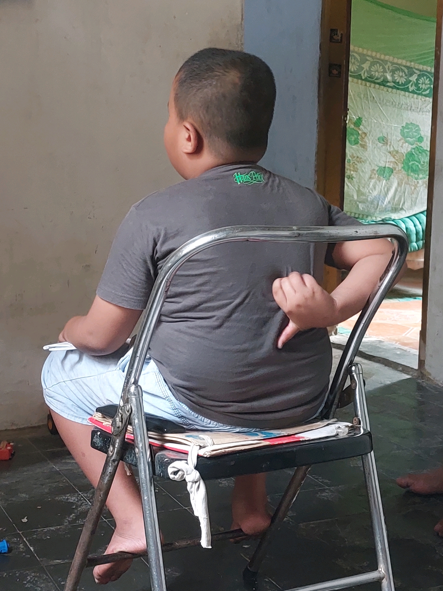A young boy, viewed from the back and side, sits on a metal folding chair in what appears to be an indoor setting. He is wearing a dark grey t-shirt with a green logo on the upper back and light blue shorts. His right arm is bent, with his hand positioned behind his back, forming a thumbs-down gesture. He is holding a white object, possibly a pen or pencil, in his left hand. The chair has a black seat with some papers or books underneath it, and a white cloth tied to the back support. The floor is tiled with dark grey tiles. In the background, a green curtain with a floral pattern hangs in an open doorway. The wall on the left is plain and grey. There are hints of toys on the floor to the left and a bare foot visible on the right side of the frame. This scene is located in Sarangan, Indonesia.