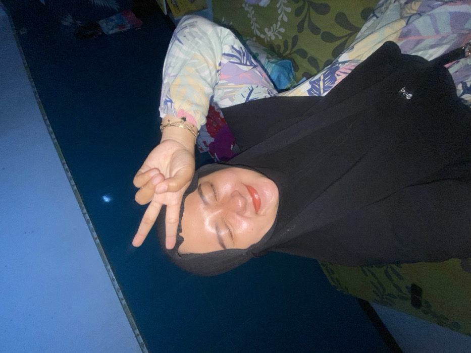 A woman wearing a black hijab and patterned pajamas lies on a green and white patterned surface. She is facing the camera, smiling with her eyes closed. Her right hand is raised, making a peace sign. The background includes a blue surface and what appears to be furniture. The image is dimly lit, giving it a somewhat dark and casual feel.