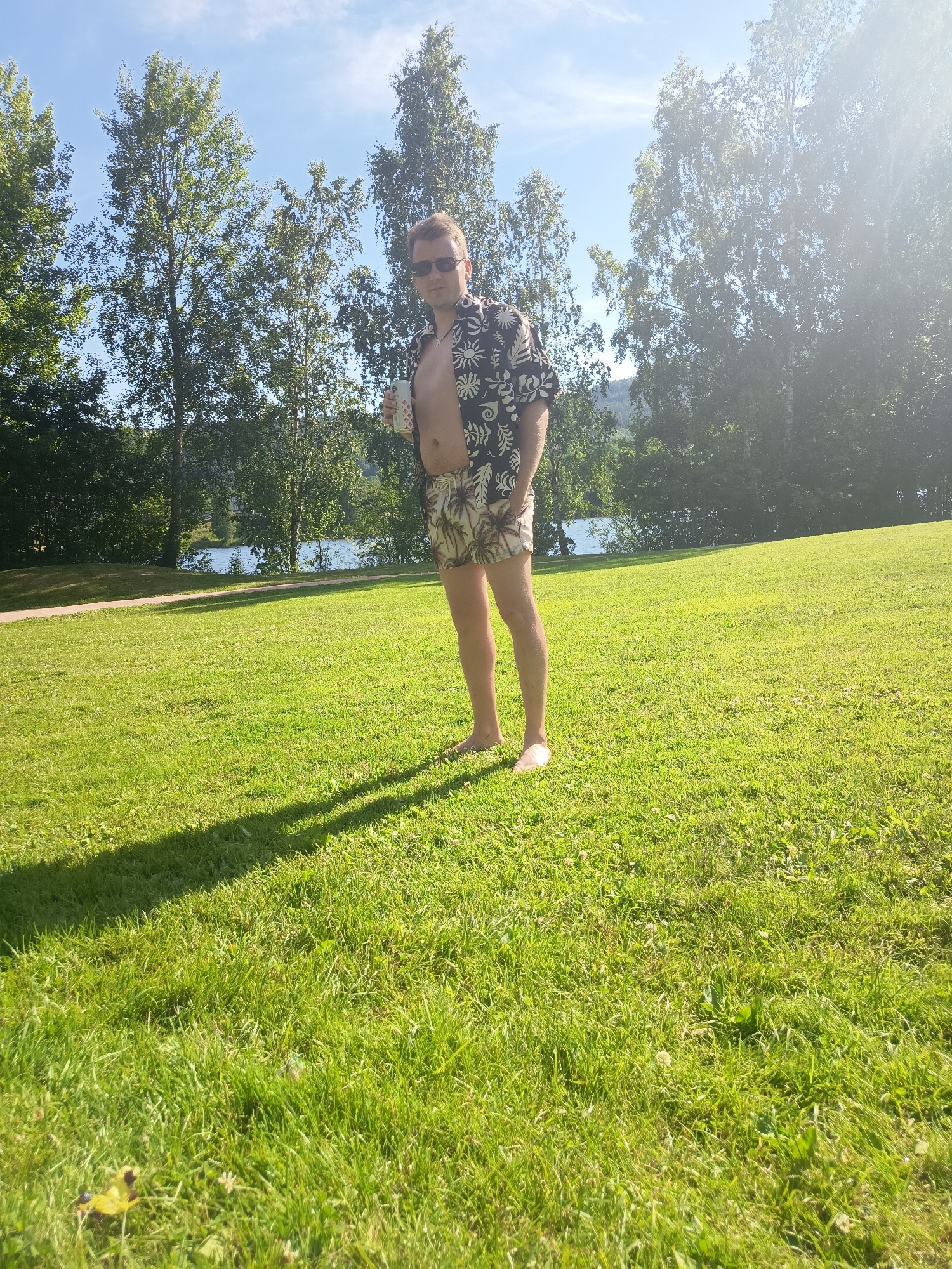 A man stands in a grassy park, holding a beverage can. He is wearing a black shirt with a white floral pattern, patterned shorts, and sunglasses. The man is barefoot. Behind him, trees and a body of water are visible under a bright, sunny sky. The scene is an outdoor park in an unspecified location.