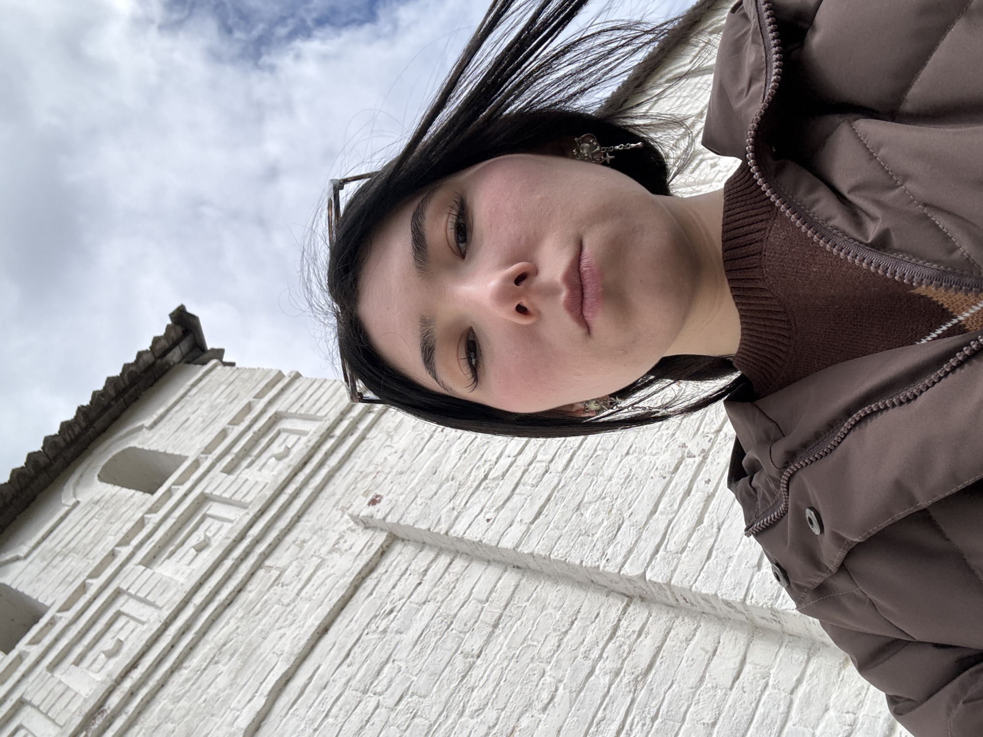 A young woman stands in Sviyazhsk, Russia. She is facing the camera, holding it at a high angle. She has dark hair, glasses perched on her head, and is wearing a brown puffer jacket over a brown sweater. She is wearing earrings. Behind her is a white brick building with decorative brickwork. The sky is overcast.