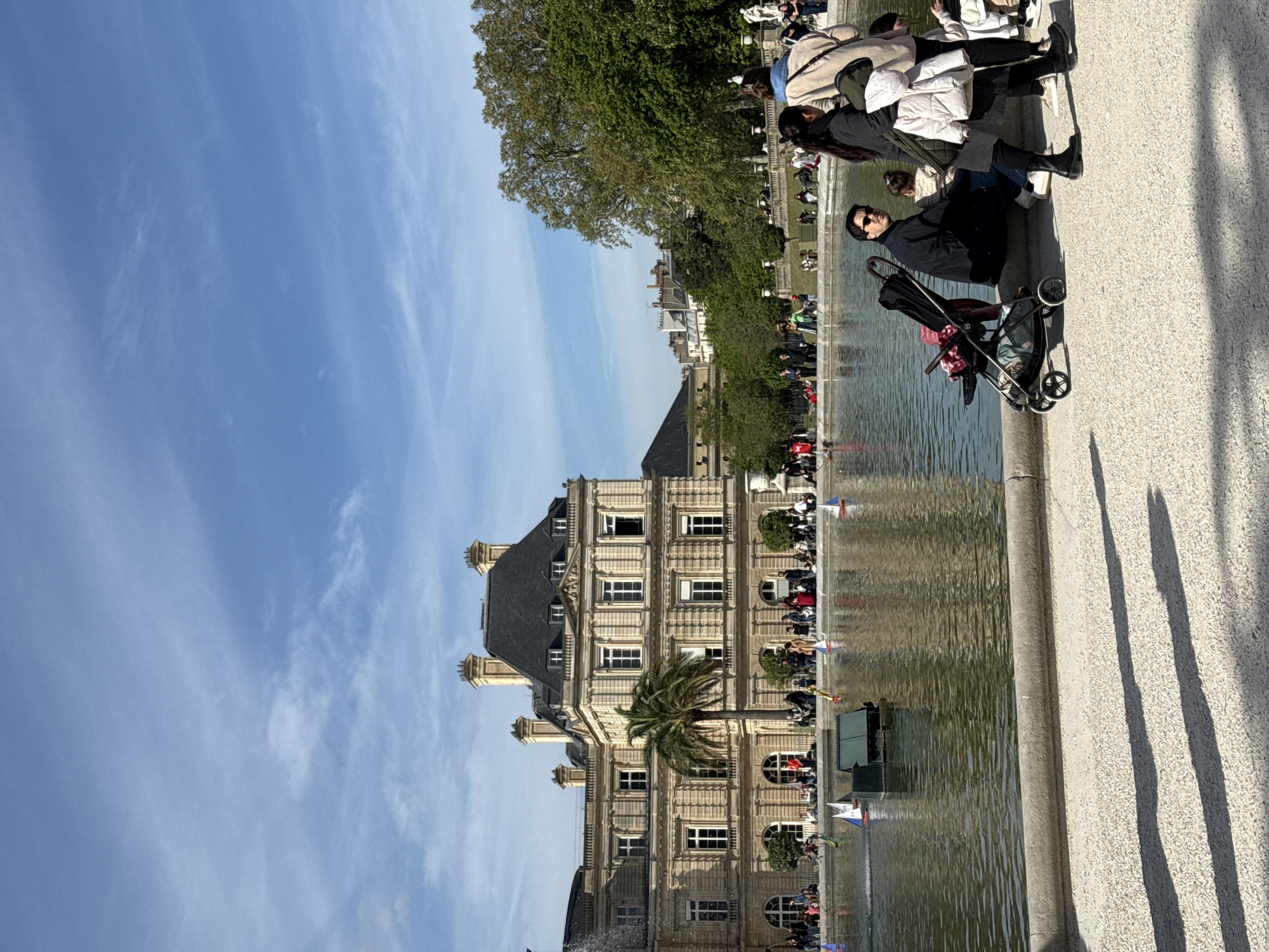 The image depicts the Luxembourg Palace and its surrounding garden in Paris, France. The palace, a multi-story building with symmetrical windows and a dark roof, is centrally framed. In front of it is a large rectangular pool of water, which reflects the palace's facade. People are situated along the edge of the pool, and some are operating small sailboats. A man with sunglasses is standing next to a baby stroller. Additional people are sitting and standing behind him and farther along the pool's edge. A tree stands next to the palace. The sky is blue, with scattered clouds. The ground is a light-colored concrete surface.