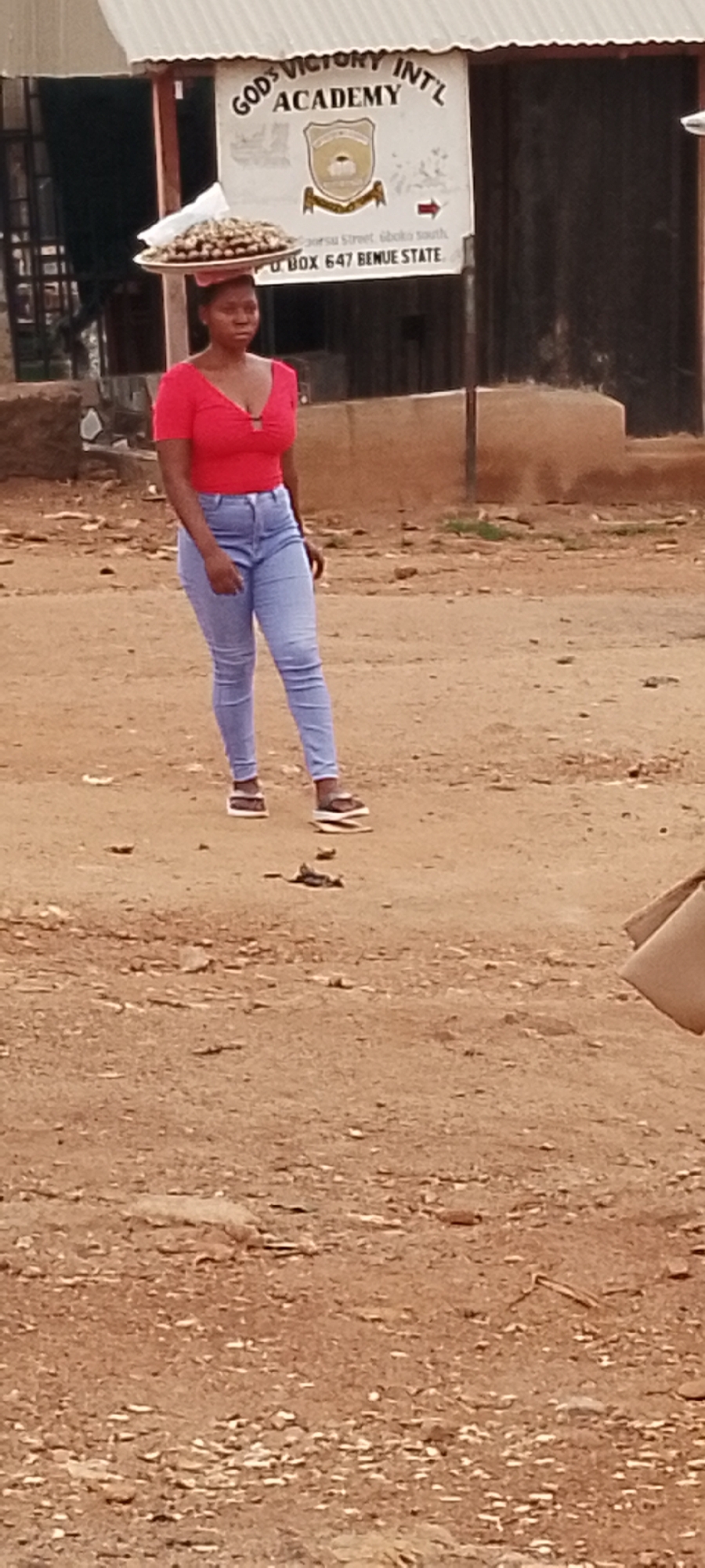A full-body, vertical shot shows a young Black woman standing on a dirt ground in Gboko, Nigeria. She is wearing a red V-neck crop top and light blue, high-waisted jeans, and white sandals. Her hair is pulled back in a bun. On her head, she carries a large, round, metal tray piled high with what appears to be nuts or small fruits. Behind her, a sign is visible, reading "GOD'S VICTORY INT'L ACADEMY" and "P.O BOX 647 BENUE STATE." The environment is dusty and appears to be an outdoor, possibly unpaved, street or market area, with some scattered debris on the ground. The lighting suggests it is daytime.