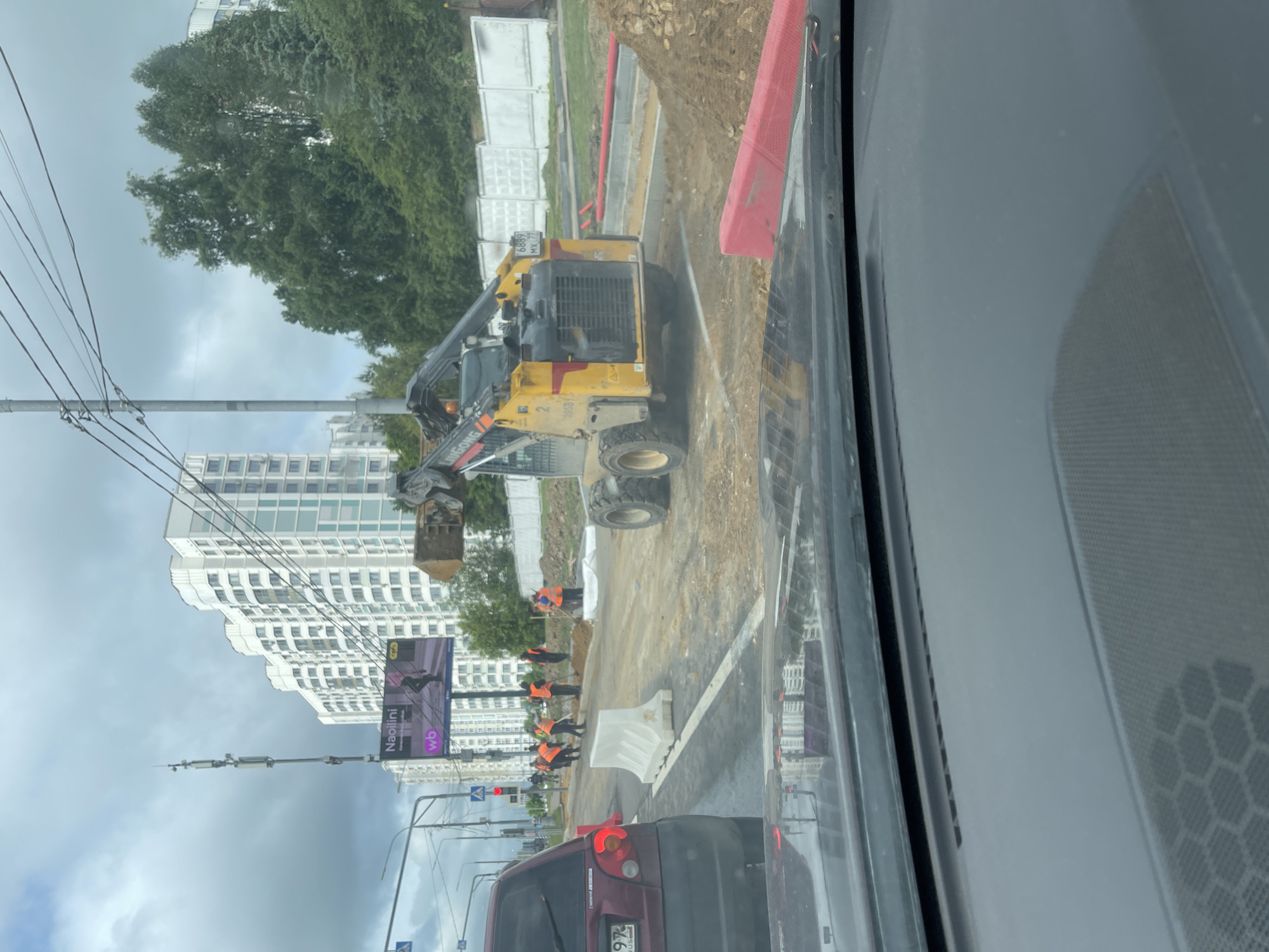 The image shows a construction scene in an urban environment. A yellow skid-steer loader is in the foreground, seemingly engaged in moving earth or materials. Construction workers in orange vests are visible, apparently working on the road or sidewalk. A red car is present on the road, suggesting traffic is active nearby. In the background, there are residential buildings and a billboard for "Naoiini". The number plate of the red vehicle indicates that it is in Moscow, Russia. Overhead power lines are noticeable, adding to the urban landscape.