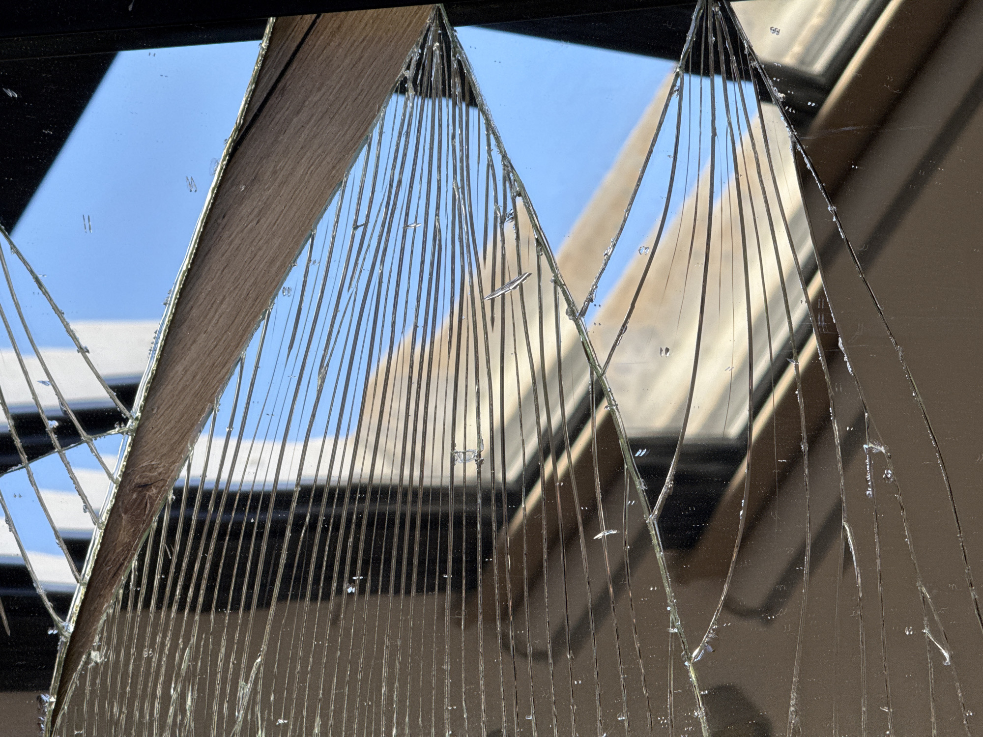 The image presents a close-up view of a severely shattered pane of glass or a mirror, displaying intricate patterns of cracks. The breakage has created numerous sharp, irregular fragments and long, slender shards that radiate outwards from several points. Many of these cracks run predominantly vertically, forming a dense, linear texture across large sections of the glass.
Through the clear gaps and reflected surfaces of the broken glass, a bright, clear blue sky is visible, particularly in the upper left quadrant, indicating a sunny day. Reflections of urban architecture, such as parts of buildings or structures, are also discernible within the fragmented surfaces, suggesting an outdoor or semi-outdoor setting. A distinct wooden element, possibly a frame, a beam, or part of a background structure, intersects the shattered glass diagonally from the upper left, adding a contrasting texture.
The scene captures a moment of damage or destruction, yet the resulting patterns of the broken glass create an abstract and visually arresting composition. The bright lighting and reflections are consistent with daylight hours. The specific location is Tel Aviv, Israel, though no distinct landmarks are visible to confirm this directly. There is no explicit activity taking place, nor is any text visible in the frame.