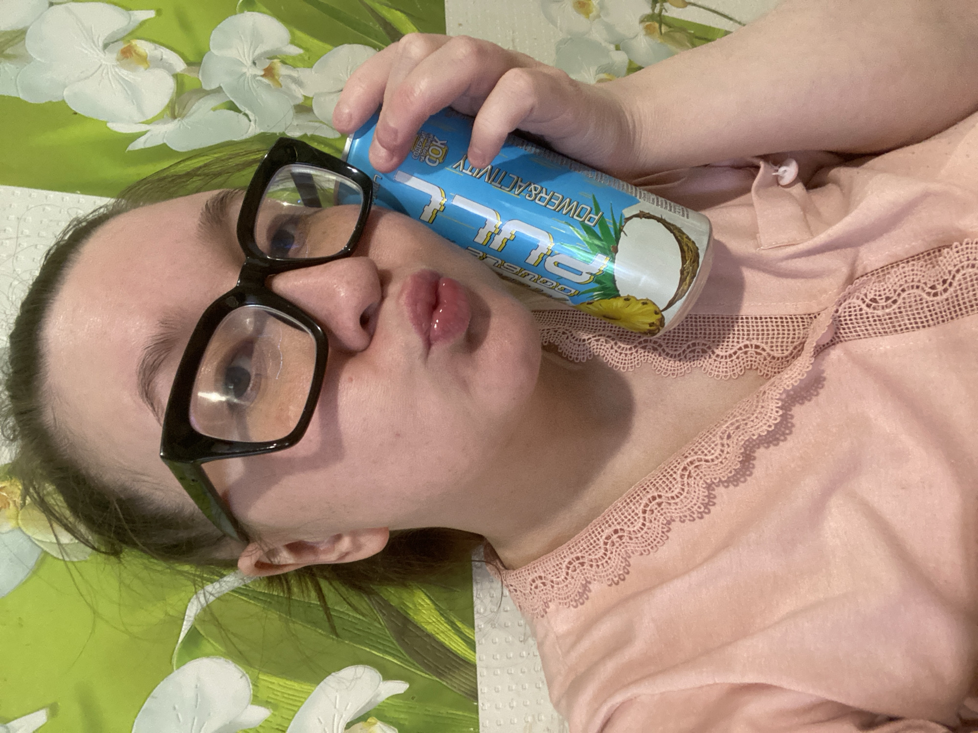 A close-up image shows an adult individual, likely female, indoors in the city of Chaikovsky, Russia. The subject has light skin, dark hair, and wears black, thick-framed eyeglasses. A light pink garment with a scalloped lace trim along a V-neckline covers the upper torso. The subject's lips are pursed forward. A light blue aluminum beverage can, featuring illustrations of a pineapple and a split coconut, is held in the subject's right hand. Visible text on the can includes "DOUBLE RICH," "JUICE," and "POWER & ACTIVITY" in English and Cyrillic. The background consists of a vibrant lime green wallpaper patterned with multiple white orchid flowers, some exhibiting subtle yellow centers. A textured, light-colored horizontal border is visible below the floral wallpaper.