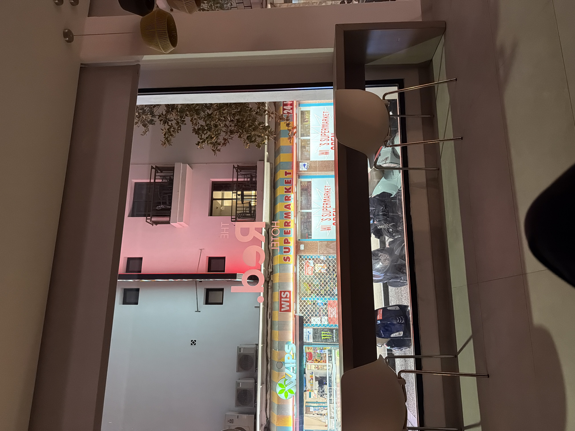 The image shows a view through a storefront window. The storefront is for "WI'S SUPERMARKET" and "24H FRESH FRU", and also displays the word "APPL". The exterior features a colorful striped facade above the supermarket entrance. Inside the supermarket, various products are visible, including what appears to be a display of Monster Energy drinks.
To the left of the supermarket, a building with a pink illuminated facade is visible, featuring the text "THE" and "b.o.b". There are also several windows and air conditioning units on this building.
Outside the supermarket, several motorcycles are parked. The scene appears to be set at dusk or night, with artificial lighting illuminating the storefront and the adjacent building. The overall setting suggests an urban environment, likely a commercial street in Sant Antoni De Portmany, Spain, given the location context. There are no people clearly visible in the image. The mood is that of a vibrant, well-lit commercial area at night.