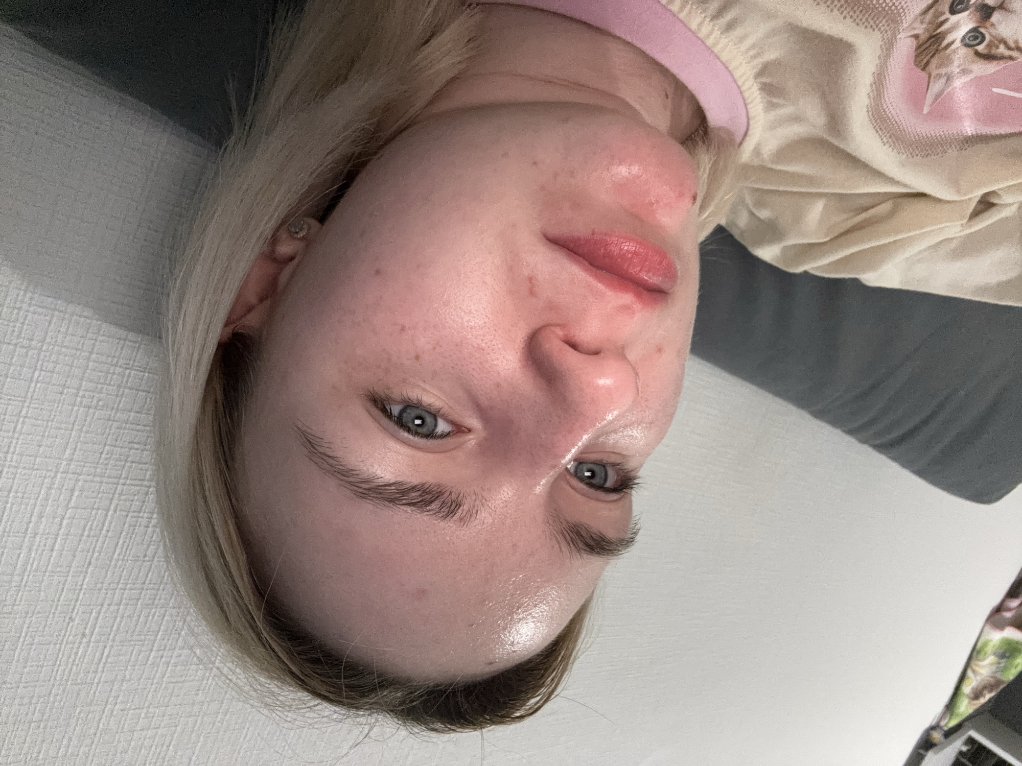 A close-up shot of a fair-skinned person with blue eyes and blonde hair, angled so that their face is the primary focus. The person is wearing a light-colored top with a pink trim and a cat design. The background includes a gray, textured surface, likely a couch or other soft furnishing, and a plain white wall. There is no indication of the image being located in Vsevolozhsk, Russia.