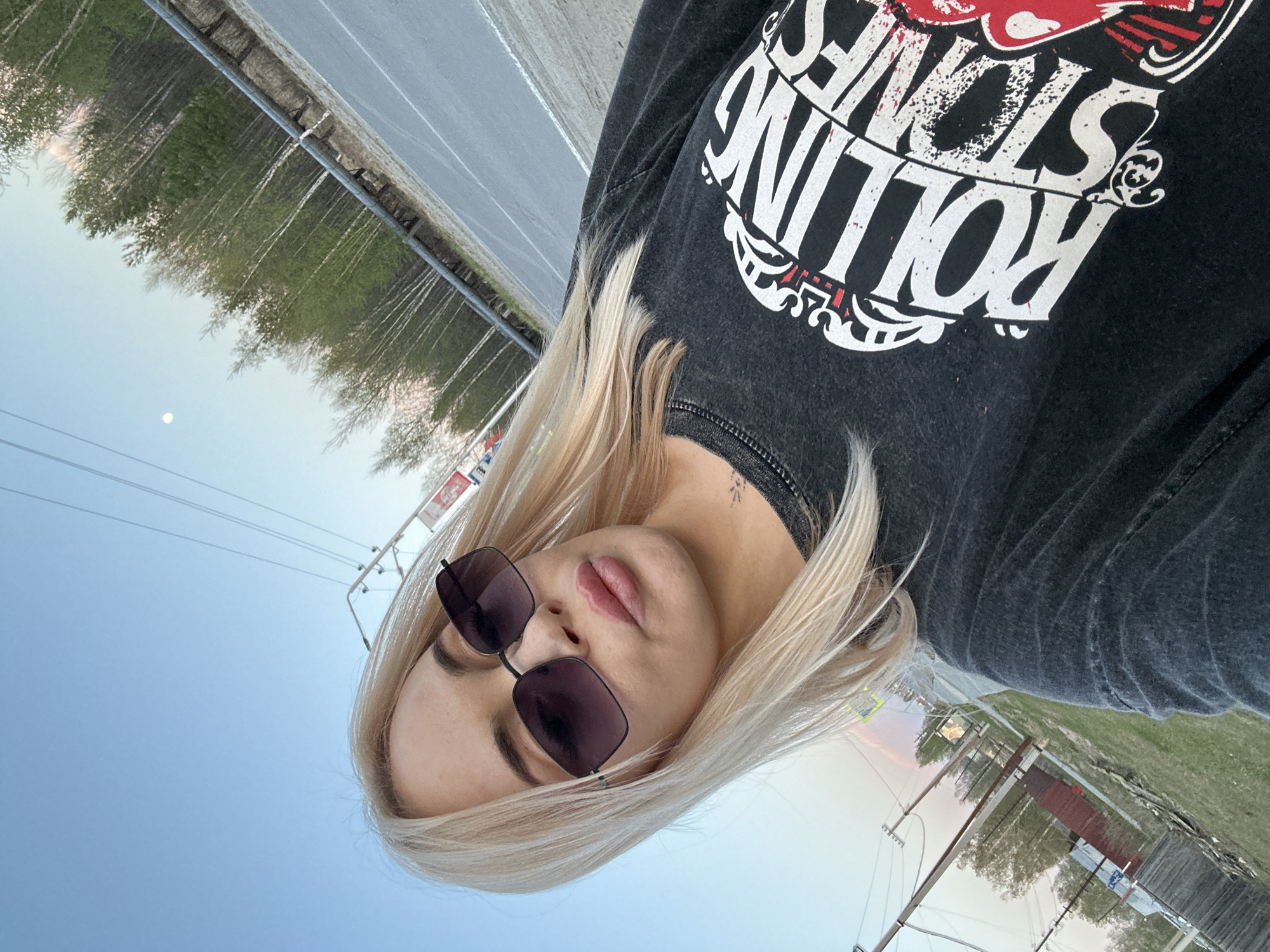 A woman with blonde hair and sunglasses is wearing a black "Rolling Stones" shirt. The photo appears to be taken outdoors in Высокий, Russia. In the background, there is a road, trees, a utility pole, and the sky.