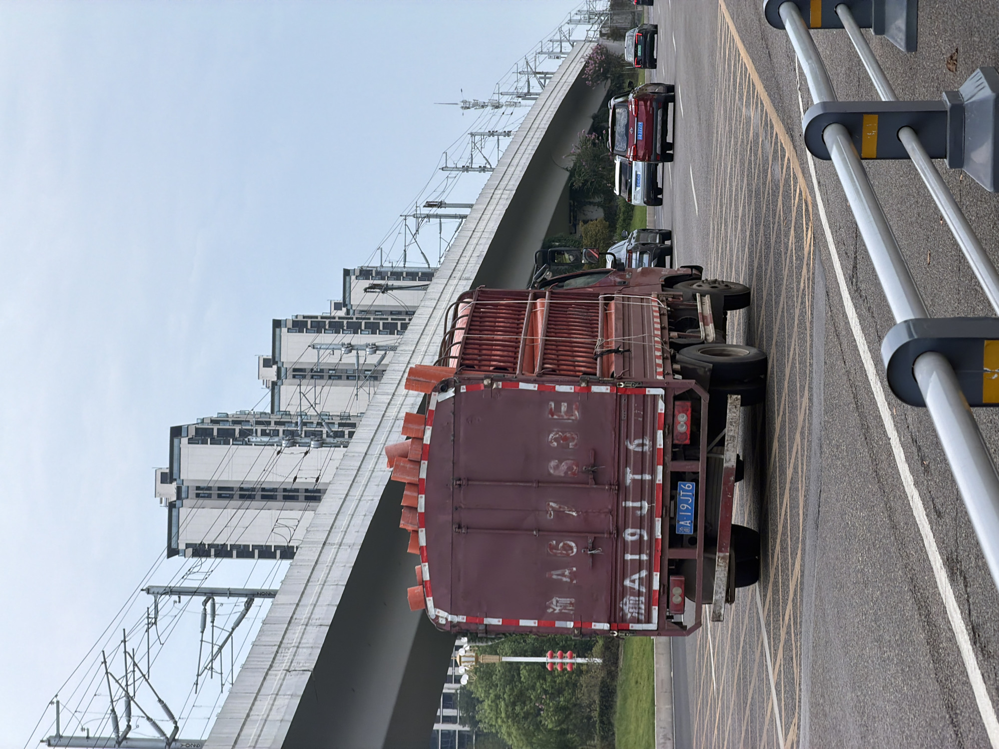 In the city of Jiangjin, China, a red truck is driving on a road under an elevated highway. The truck is carrying an orange object on its roof. Several cars are also on the road, driving in the same direction as the truck. In the background, there are tall buildings and a sky with clouds. The scene appears to be an urban environment with a focus on transportation infrastructure.