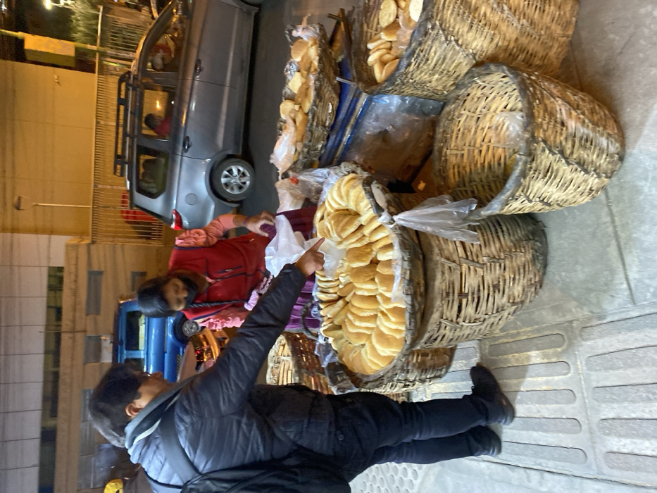 This image captures a street vending scene in an urban area of La Paz, Bolivia, during low-light conditions, likely evening or early morning. In the foreground, on a concrete sidewalk featuring tactile paving strips, a male customer interacts with a female bread vendor. The customer, dressed in a dark jacket, dark pants, and a dark backpack, is shown in profile pointing his right index finger at a large, round wicker basket filled with dozens of light-colored bread rolls arranged in concentric circles. The female vendor, positioned behind the bread display, wears a red vest over a pink-ruffled blouse, a dark skirt, and a black protective face mask; her dark hair is styled in braids. She holds a white plastic bag in her left hand, indicating an active transaction. Multiple additional wicker baskets containing similar bread items are arranged around the main display, some elevated on what appears to be a metal stand. In the background, on the adjacent street, a blue pickup truck and a grey SUV with a roof rack are parked alongside indistinct building facades, all under artificial illumination.