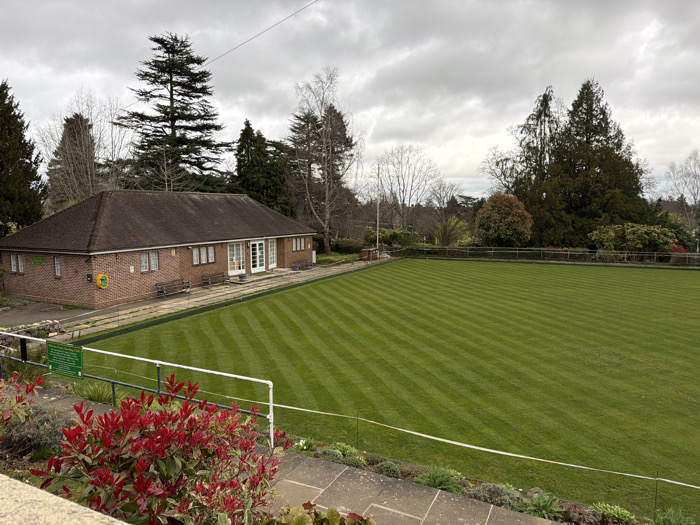 An outdoor scene captures a bowling green facility in Malvern Hills, United Kingdom, under an overcast sky. In the foreground, vibrant red-leafed shrubs are visible above a concrete retaining wall, topped by a white metal railing. The central midground features a large, rectangular, unoccupied bowling green, meticulously mown with distinct alternating light and dark green stripes. A narrow paved perimeter borders the green. To the left, a single-story brick clubhouse with a brown hip roof, white-framed windows, and white double doors faces the green. Four dark benches are positioned on the paved area directly in front of the clubhouse. A circular yellow sign with black text and a rectangular green informational sign are affixed to the clubhouse wall and the railing, respectively. The background consists of a dense arrangement of mature trees, including tall conifers and deciduous trees with bare branches, forming a natural backdrop. No individuals are present, and no active play is occurring on the green.