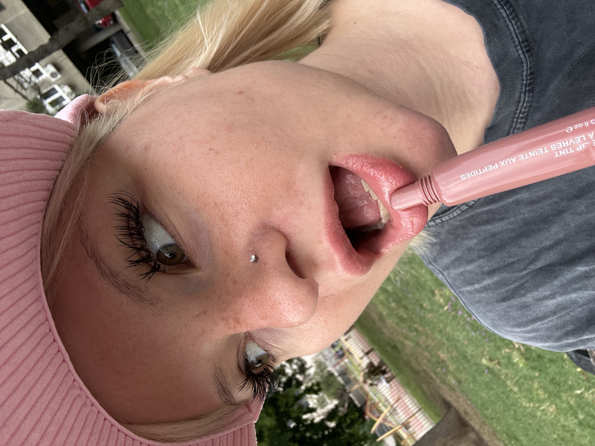 This is a close-up, low-angle, eye-level shot of a young person's face, tilted to the right, with a blurred background of green grass and what appears to be a fence or playground structure. The person has blonde hair, fair skin with freckles, and is wearing a pink beanie. Their eyes are a light brown, and they have long, dark eyelashes. A small, silver stud piercing is visible on the person's nose. Their mouth is open, revealing teeth and tongue, and they are holding a light pink lip tint tube with text on it. The lip tint appears to be applied to their lips. The person is wearing a dark grey denim top. The scene is outdoors, likely during the daytime, with soft lighting suggesting it could be overcast or late afternoon. The overall mood is casual and playful, with the focus on the facial features and the lip product. Visible text on the lip tint tube reads "LIP TINT A LEVRES TEINTE AUX PEPTIDES .3 fl oz e".