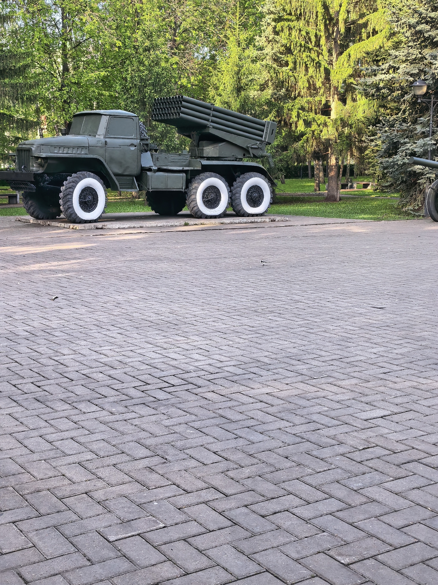 The image depicts a BM-21 Grad multiple rocket launcher mounted on a Ural-375D truck chassis, located in Sterlitamak, Russia. The vehicle is painted green and features distinctive whitewall tires. It is positioned on a paved area constructed of gray herringbone-patterned bricks. The background consists of various deciduous and evergreen trees, suggesting a park or memorial setting. Another cannon is partially visible on the right side. The scene appears to be a static display of military equipment. There are no people visible in the image.