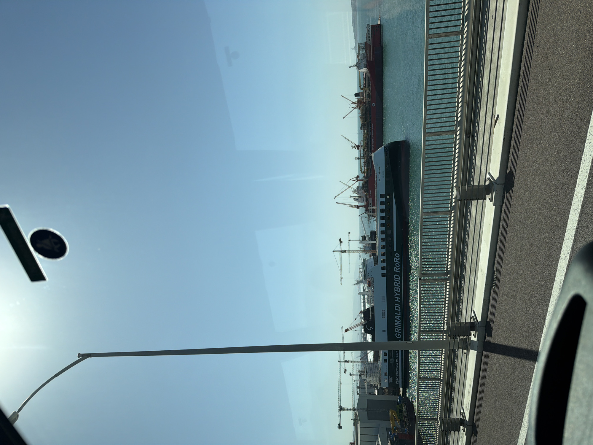 The image shows a harbor scene viewed from a high vantage point, possibly a bridge or elevated road, in Altınova, Turkey. Several cargo ships, including one with "GRIMALDI HYBRID RORO" painted on its side, are docked in the harbor. Numerous cranes are visible, indicating active port operations. The sky is clear blue. A section of the bridge or road, with its guardrail and surface, is in the foreground. Reflections are visible on the window through which the photo was taken.