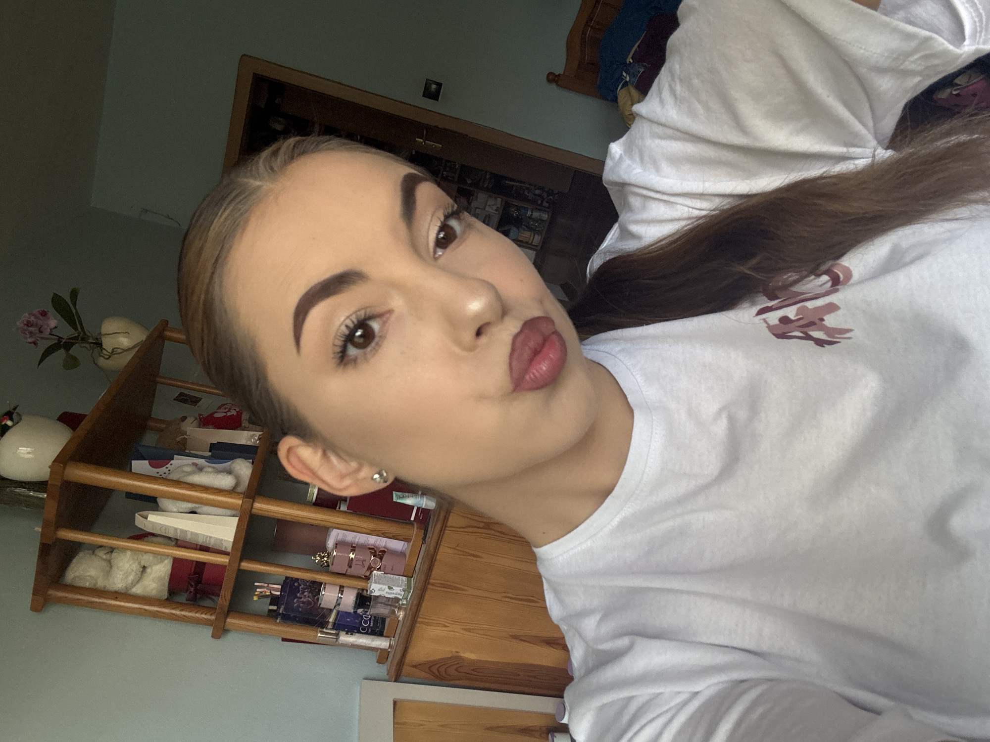 A young Caucasian woman is taking a selfie indoors. She is wearing a white t-shirt and has her hair pulled back into a ponytail. Her facial expression is one of a pout. Behind her, a wooden shelf unit holds various items, including teddy bears and perfume bottles. To the right, a doorway leads to another room with a bed. The walls are painted a light blue and a muted green color. The lighting suggests it is daytime, possibly with natural light coming from a window. The setting appears to be a residential interior. The location cannot be determined to be Sofia, Bulgaria from the image content.