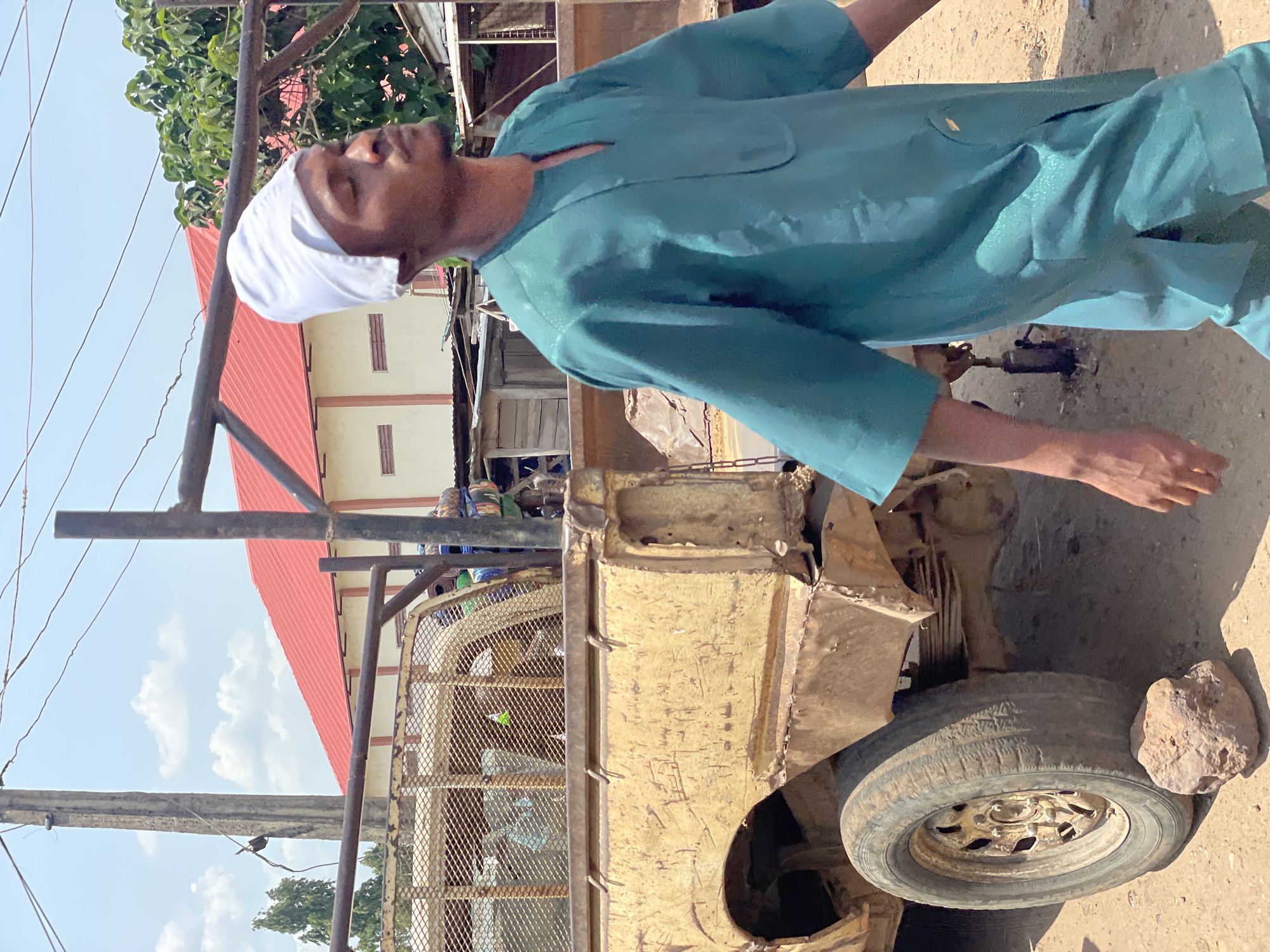 The image depicts a man wearing a light teal shirt and a white head covering standing next to a heavily damaged, yellow vehicle. The vehicle appears to be a truck, with a flat tire propped up by a rock. The background shows a building with a red roof and light-colored walls. Utility poles and wires are also visible in the sky. The location appears to be an urban or semi-urban area.