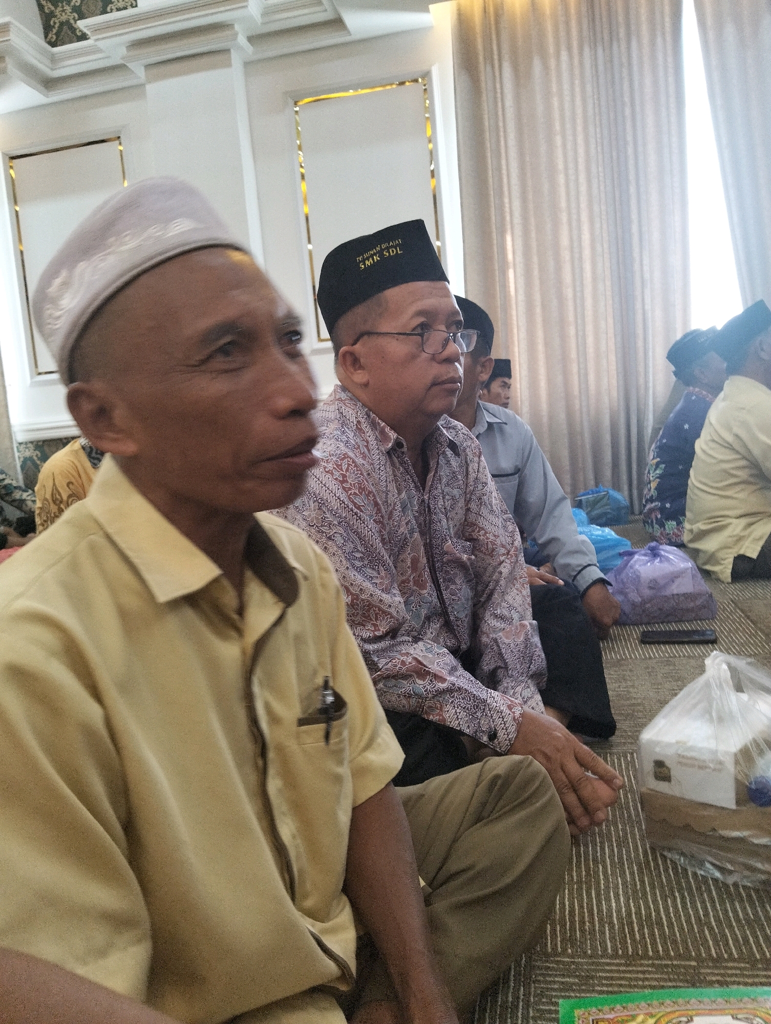 The image shows a group of men sitting on the floor in a room. The man in the foreground is wearing a light yellow short-sleeved collared shirt and a white cap. He is looking to the right. Another man, slightly behind and to the right of the first, is wearing a patterned shirt and a black cap with white lettering that reads "SMK SDL". He is wearing glasses and looking forward. In the background, other men are visible, some wearing caps. The room appears to be an indoor space, possibly a mosque or community hall, with ornate ceilings and patterned carpet. Several plastic bags containing items are placed on the floor. The overall scene suggests a gathering or event taking place in Dagan, Indonesia.