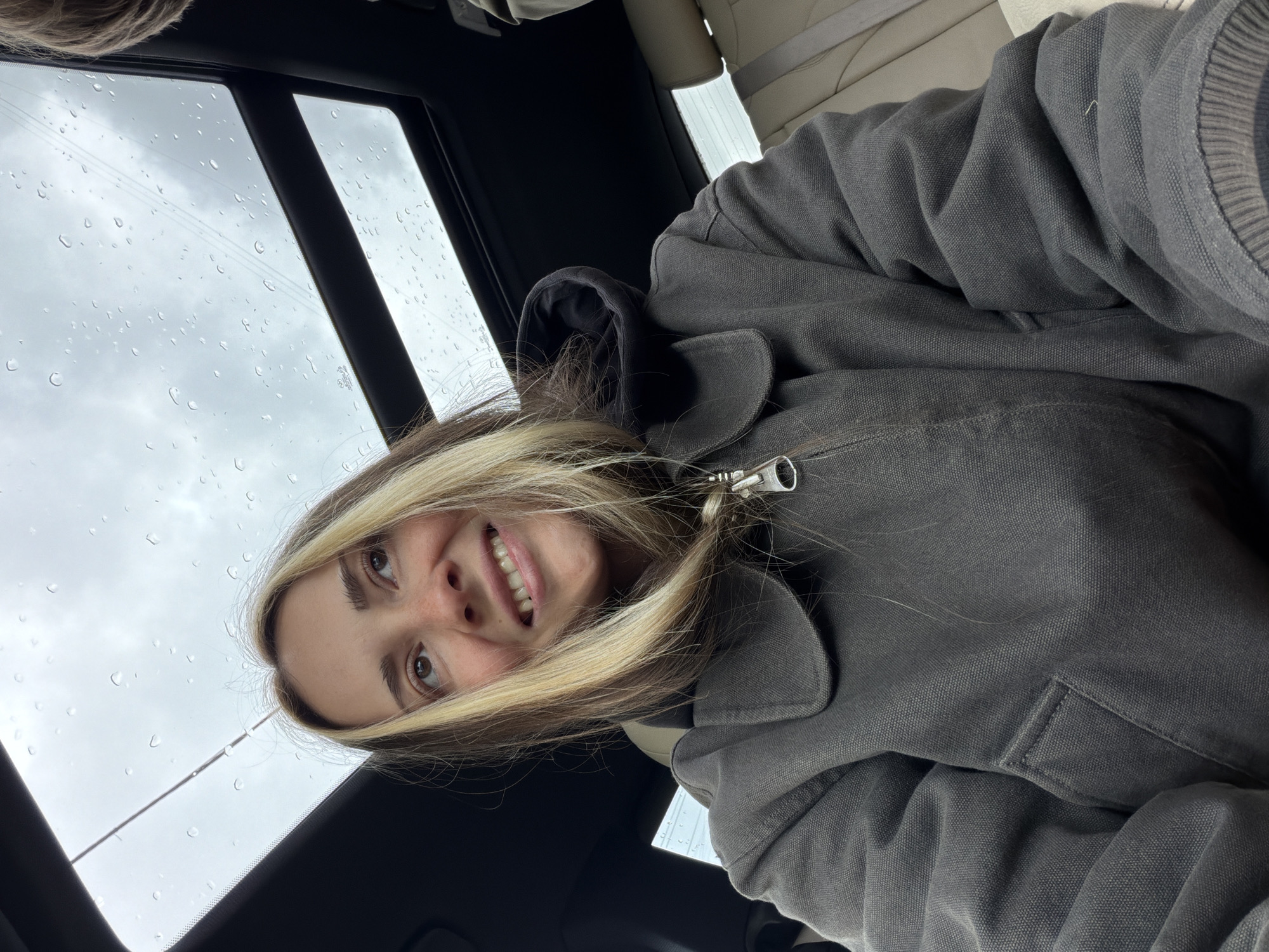 A close-up, portrait-oriented image shows a young woman inside a car, smiling and looking upwards towards the camera. Her face, featuring brown eyes and a wide smile, is framed by long, blonde and brown highlighted hair. She is wearing a dark grey or charcoal-colored jacket with a visible metal zipper pull near her shoulder.
The setting appears to be the interior of a vehicle, possibly a car, with light-colored upholstered seats visible in the background. Above and behind her head, a panoramic roof or sunroof window is prominent, covered with numerous raindrops. Beyond the car window, an overcast and cloudy sky is visible, indicating rainy or recently rainy weather during daytime. The overall mood conveyed by her smile is pleasant and content. There is no visible text in the image. The provided location context is Khimki, Russia.