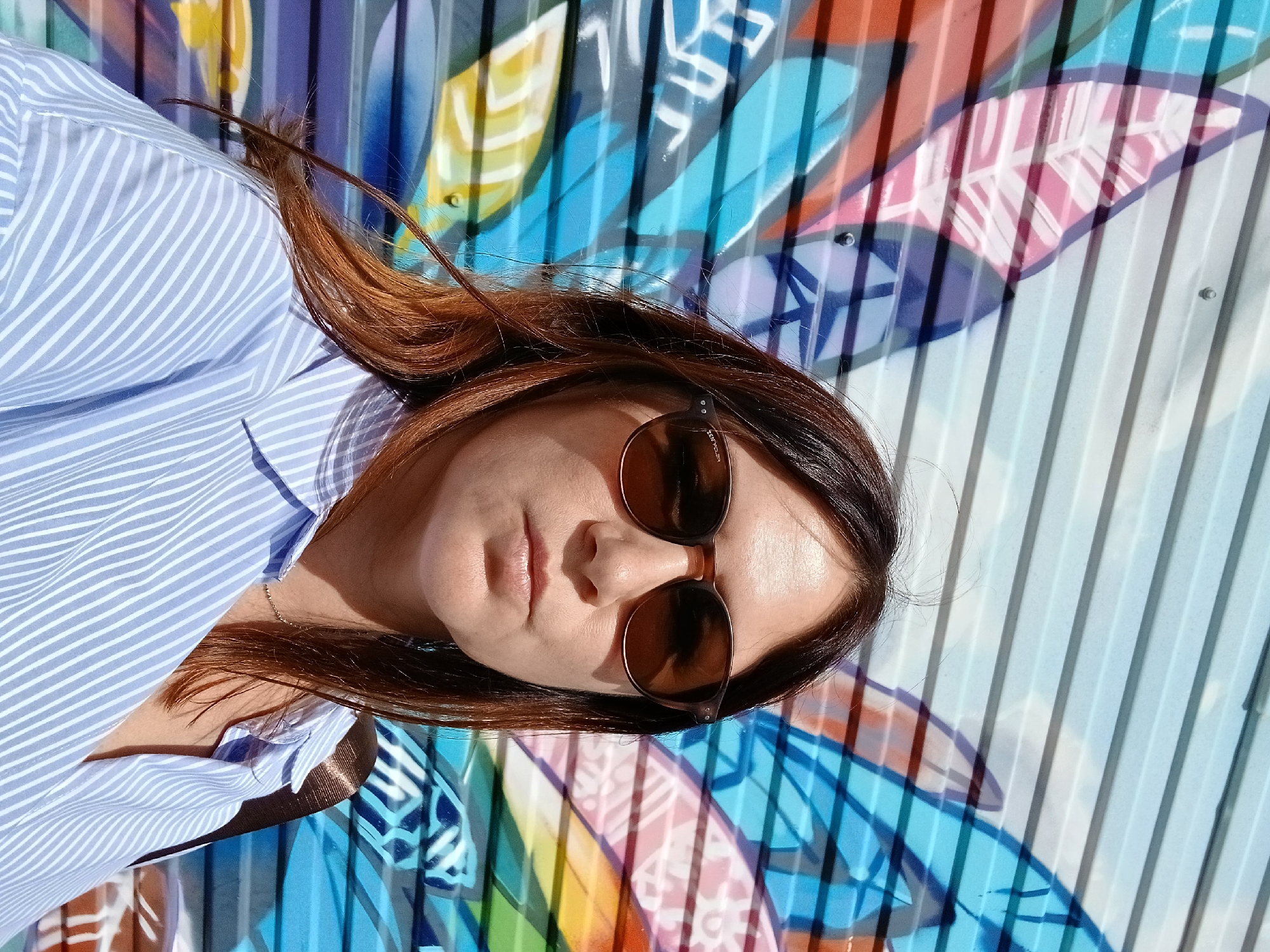 In the city of Unknown, Unknown, a woman stands in front of a graffiti-covered wall. The wall features large, colorful abstract shapes and patterns, predominantly in shades of blue, pink, orange, and yellow. The woman, with shoulder-length brown hair and wearing sunglasses, is looking directly at the camera. She is dressed in a light blue and white striped button-down shirt. The lighting suggests it is daytime, with shadows cast on the wall and the woman's face. The perspective is a medium close-up, focusing on the woman's upper body and face.