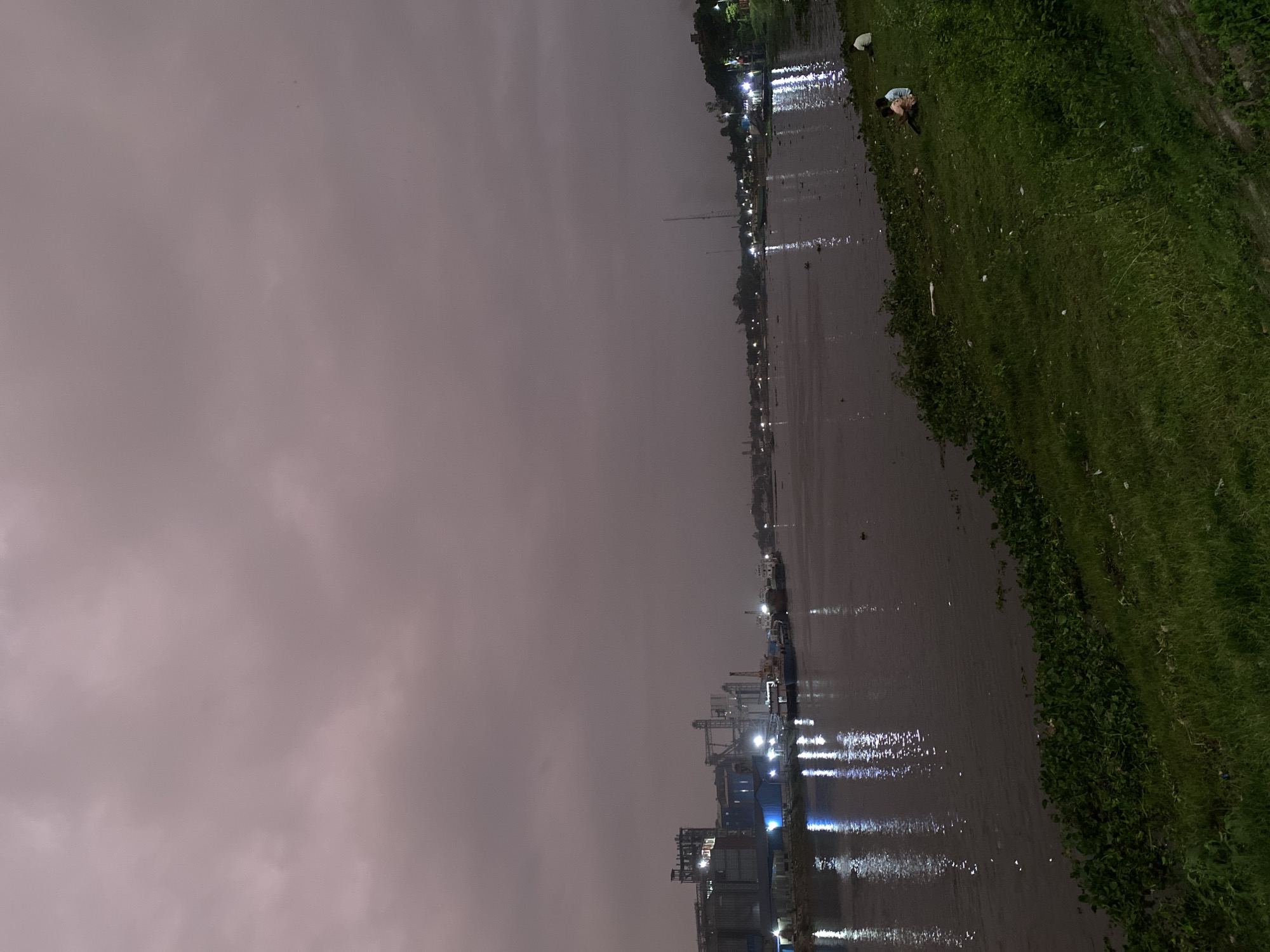 The image shows a body of water reflecting lights, likely a river or canal in Dhaka, Bangladesh, at night. The sky is overcast. On one side of the water is a grassy bank. A few people can be seen on the riverbank. The other side of the water shows buildings and structures with lights, some possibly industrial or port-related.