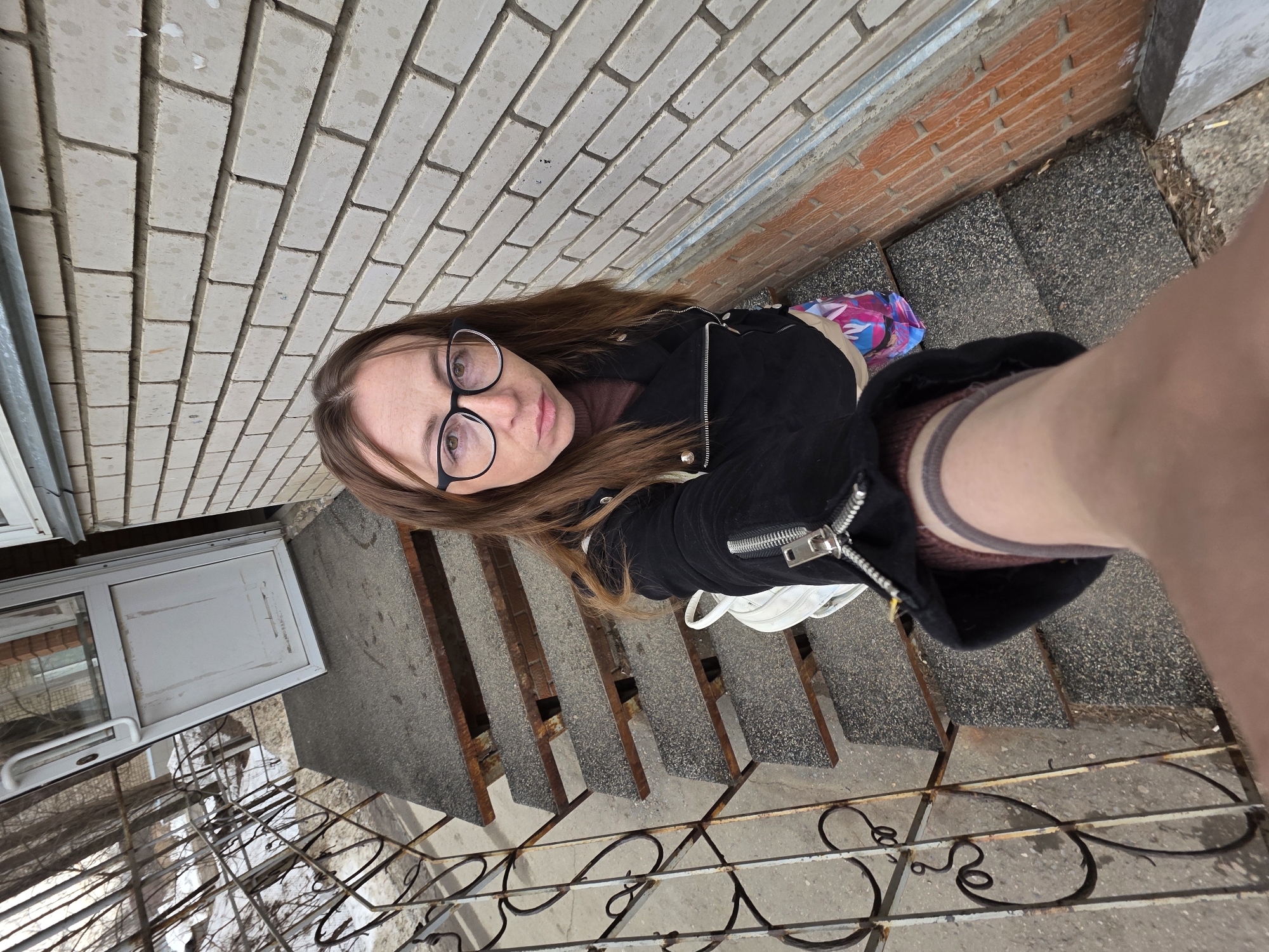 The image portrays an adult female captured in a wide-angle selfie outdoors in Chaikovsky, Russia. The subject, positioned slightly left of center, possesses shoulder-length brown hair and wears black-framed eyeglasses. She is dressed in a dark brown top, a black jacket featuring a silver zipper detail on the left chest and silver stud-like embellishments, and a multi-colored garment with pink, blue, and white patterns visible beneath the jacket. A white backpack is worn on her back, partially visible over her left shoulder. Her right arm, holding the camera, is extended into the lower right foreground of the frame.
The individual is standing on a dark grey, textured concrete staircase, with five steps clearly visible, supported by rust-colored metal structures. To her left, an aged, rusted metal railing with ornate, scrolled patterns runs parallel to the stairs. To her right, a building wall constructed from light-colored bricks, showing various dark stains, forms the immediate background. Below a lighter course of bricks, a section of reddish-brown bricks is present, separated by a horizontal silver metal trim. At the top of the staircase, a white-framed glass door with a white handle stands slightly ajar. Beyond the door and railing, a wintry or early spring outdoor environment is visible, characterized by bare deciduous trees and patches of snow on the ground. The scene is illuminated by natural daylight.