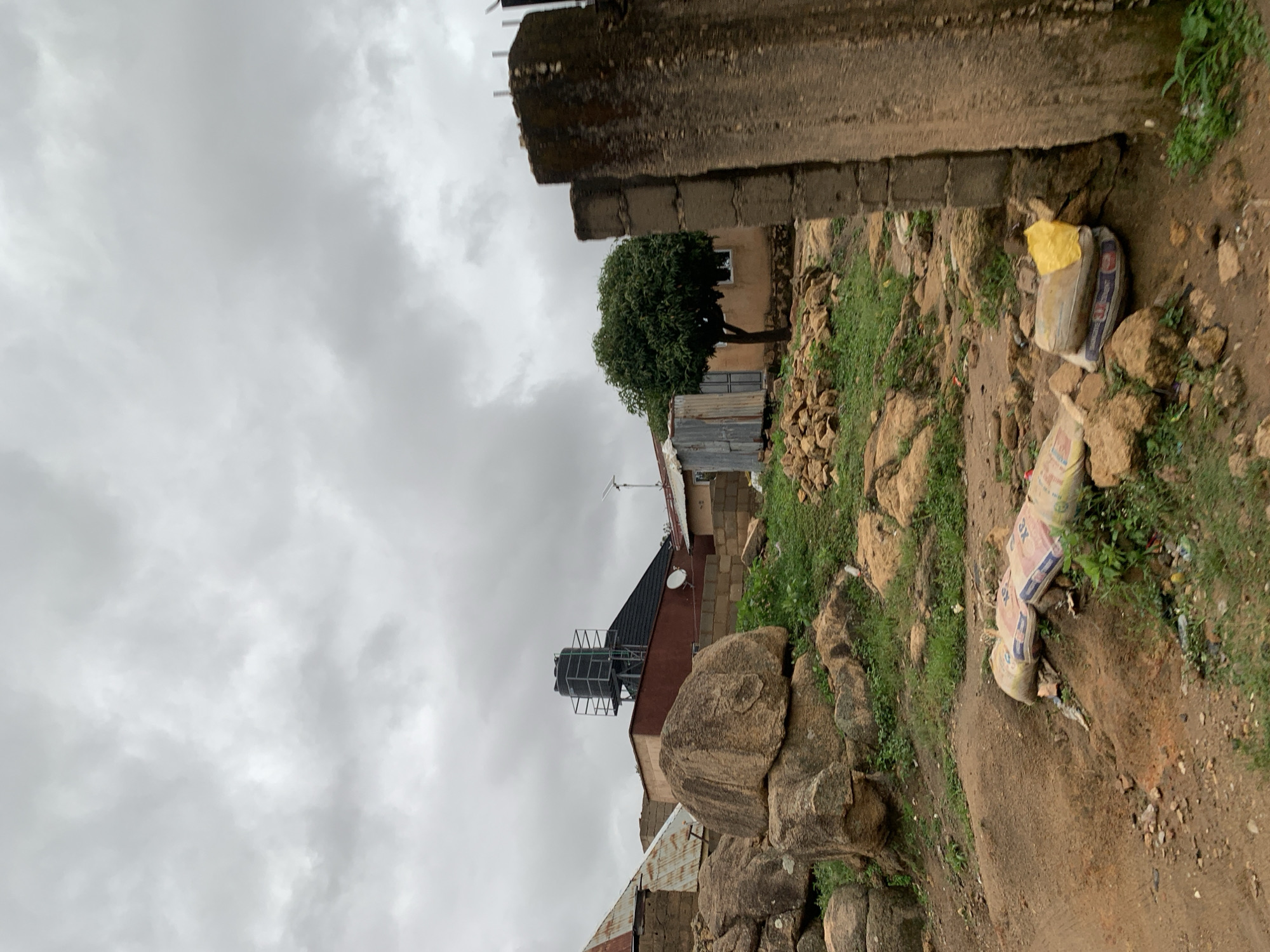 The image presents a rugged, semi-urban landscape under an overcast sky. The gray, cloudy sky dominates the upper portion, indicating a dull or humid day.
The terrain is characterized by uneven, rocky ground and dirt paths. Sparse patches of green grass and small shrubs are scattered amongst the rocks. In the foreground and extending to the right, several bags, likely containing building materials, are visible on the ground. One bag is a distinct bright yellow, while another, partly obscured, clearly shows the text "3K" and a blue and white logo.
In the midground, a cluster of simple buildings is nestled into the hilly landscape. One prominent structure features a dark, sloped roof on which a black water tank is elevated on a metal stand. A small satellite dish and what appears to be a solar panel or antenna are also mounted on this roof. Further back, another building with an earth-toned facade, possibly made of mud or adobe, has a small rectangular window. A healthy green tree with dense foliage stands in front of this building. Other structures made of corrugated metal are also present.
The scene depicts an area with basic infrastructure, likely in a developing region, consistent with its Jos, Nigeria context. No human activity is observed, and the overall impression is one of a quiet, residential or construction-adjacent area.