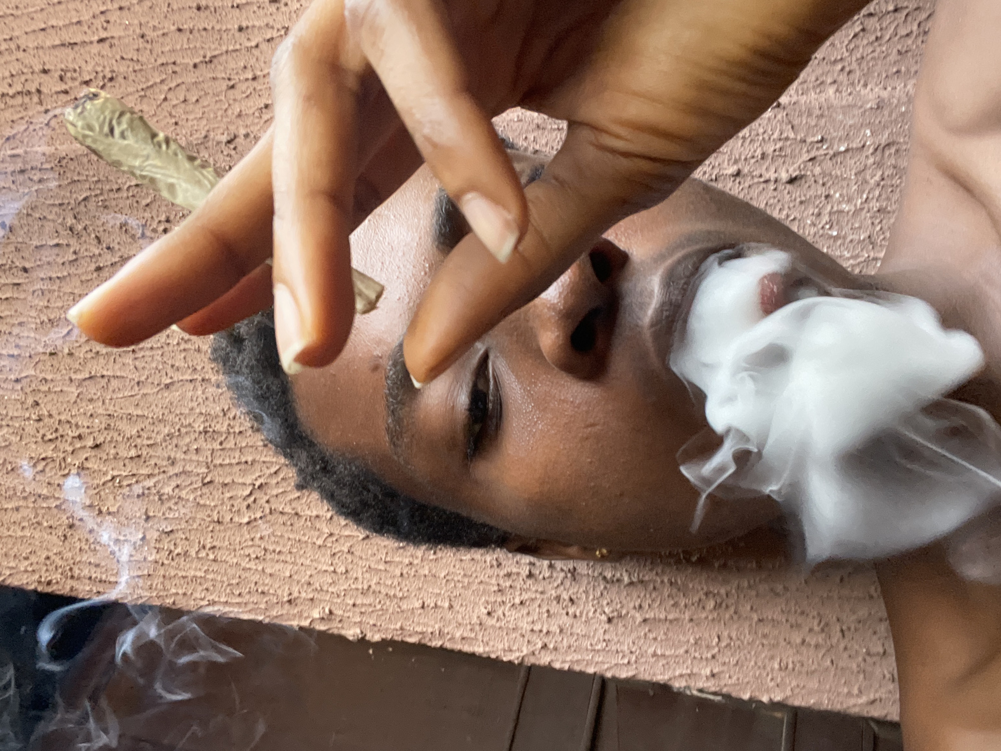 A dark-skinned person lying down exhales a large cloud of white smoke from their mouth. The person's hand holds a rolled item near their face. The background appears to be a textured surface. The person has dark hair and dark eyebrows. There is additional smoke near the bottom left of the frame. There is no indication of this image being in Onitsha, Nigeria.