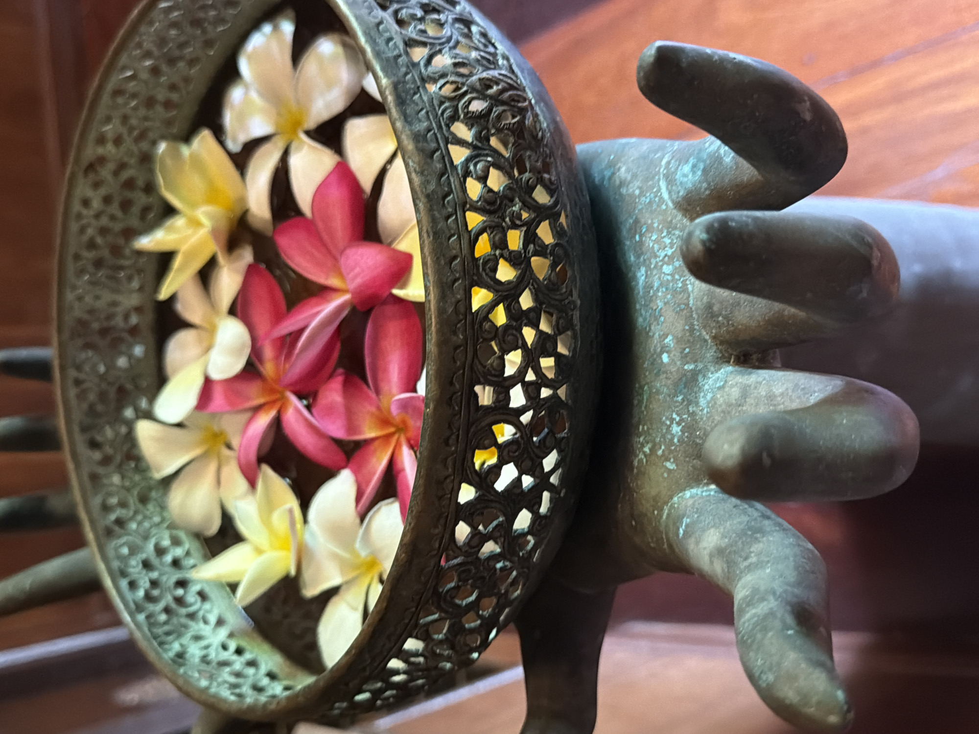 The image shows a close-up view of a decorative brass bowl filled with water and floating flowers. The bowl has an intricate, ornate pattern around its rim and is supported by a bronze sculpture of a hand. The flowers are of different colors, including white, yellow, and pink, and appear to be plumeria blossoms. The background is blurred but suggests an indoor setting with wooden flooring and possibly other decorative elements. This scene is commonly found in spa or wellness centers in tourist areas like Koh Samui, Thailand, to create a serene and aesthetically pleasing ambiance.
