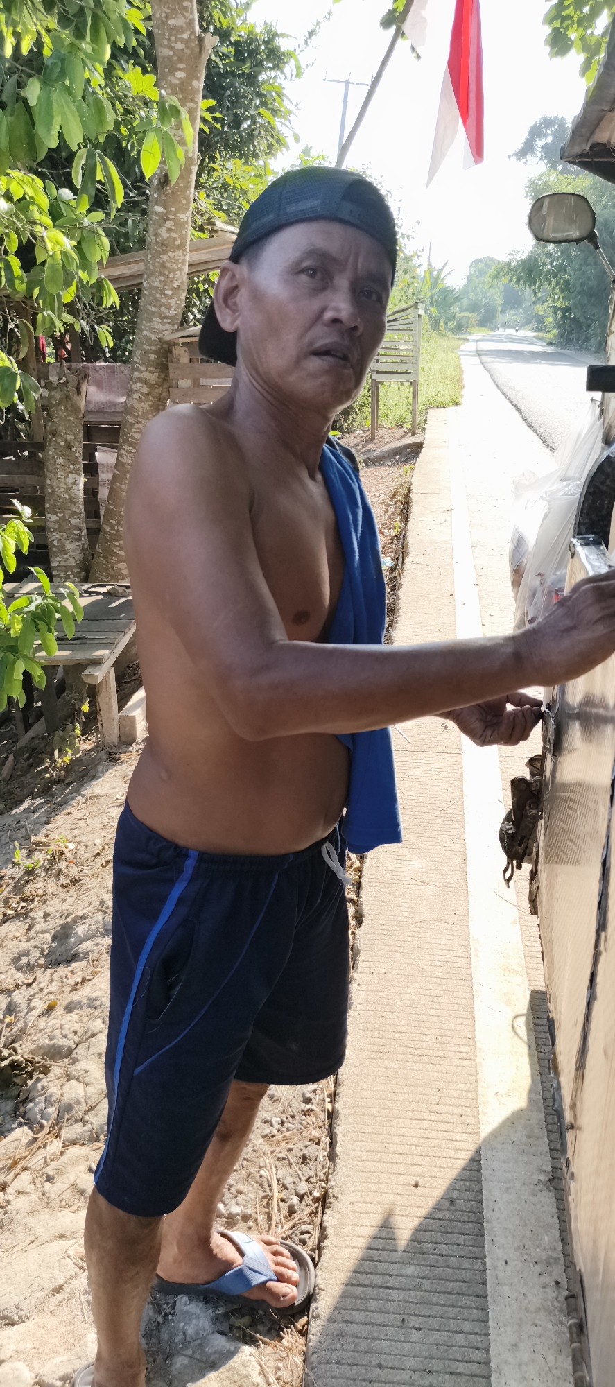 In Muaro Jambi, Indonesia, an shirtless man wearing a black baseball cap and dark blue shorts with blue trim stands next to a vehicle. A blue towel is draped over his left shoulder. He is looking towards the viewer with a slightly surprised expression and his right hand is extended, touching the side of the vehicle. The vehicle appears to be a truck or a large cargo van, with a reflective surface and some visible hardware. The man is standing on a textured concrete surface next to a dirt and gravel path. Behind him, lush green foliage and trees are visible, along with a wooden bench. Further in the background, a paved road stretches into the distance, with a few distant trees and a building. A portion of a flag, red and white, is visible hanging from a pole in the upper right corner of the image. The lighting suggests it is daytime, with the sun casting shadows.