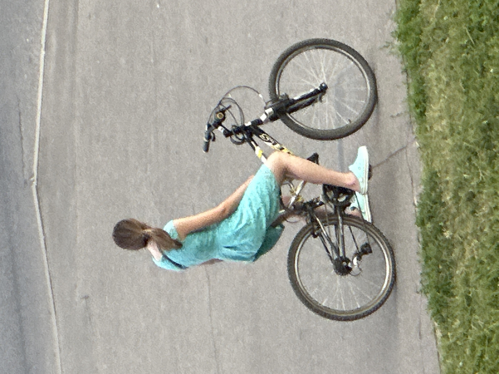 A young female with brown hair tied in a ponytail, wearing a short-sleeved turquoise dress and light blue slip-on shoes, is riding a black and yellow mountain bike. She is positioned in the middle of a paved road, with a grassy area visible in the foreground and a blurred asphalt surface in the background. The image appears to have been taken in Podolsk, Russia.