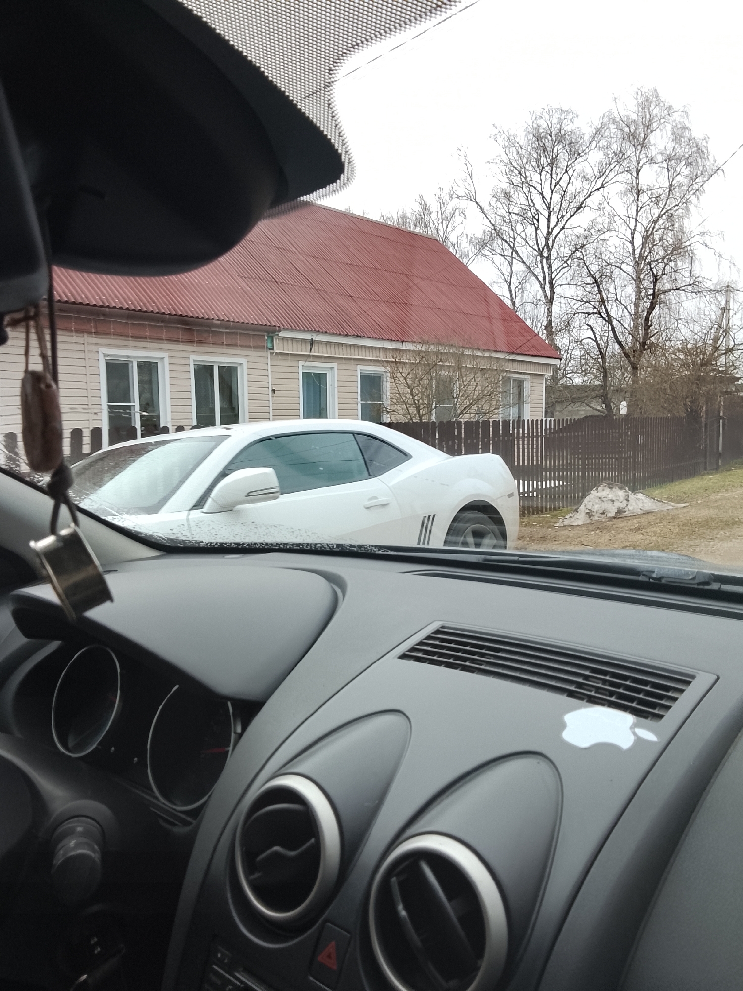 The image shows a white Chevrolet Camaro parked on a street in Высокиничи, Russia. The photo is taken from inside another car, with the dashboard and part of the windshield visible. The Camaro is parked in front of a tan single-story house with a red corrugated roof and white-framed windows. A dark wooden fence runs along the property line adjacent to the road. The sky appears overcast, and trees with bare branches are visible in the background. The dashboard of the car taking the picture is black, with circular air vents and an Apple logo sticker.