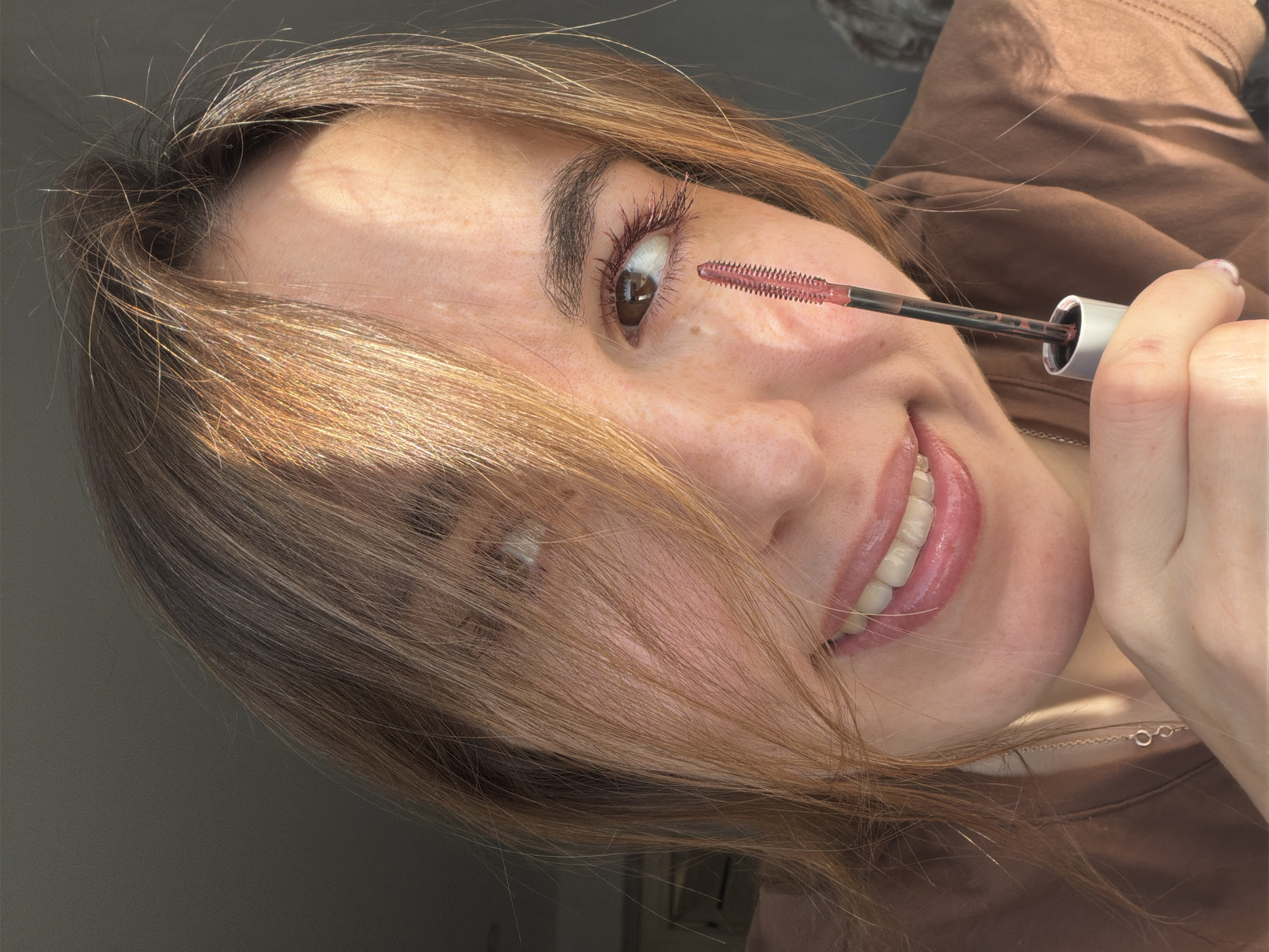 A woman with light skin and light brown hair is applying pink mascara to her upper eyelashes. She is smiling, showing her upper teeth. She is wearing a brown shirt. The location and the city of Sarapul, Russia, cannot be determined from the image.