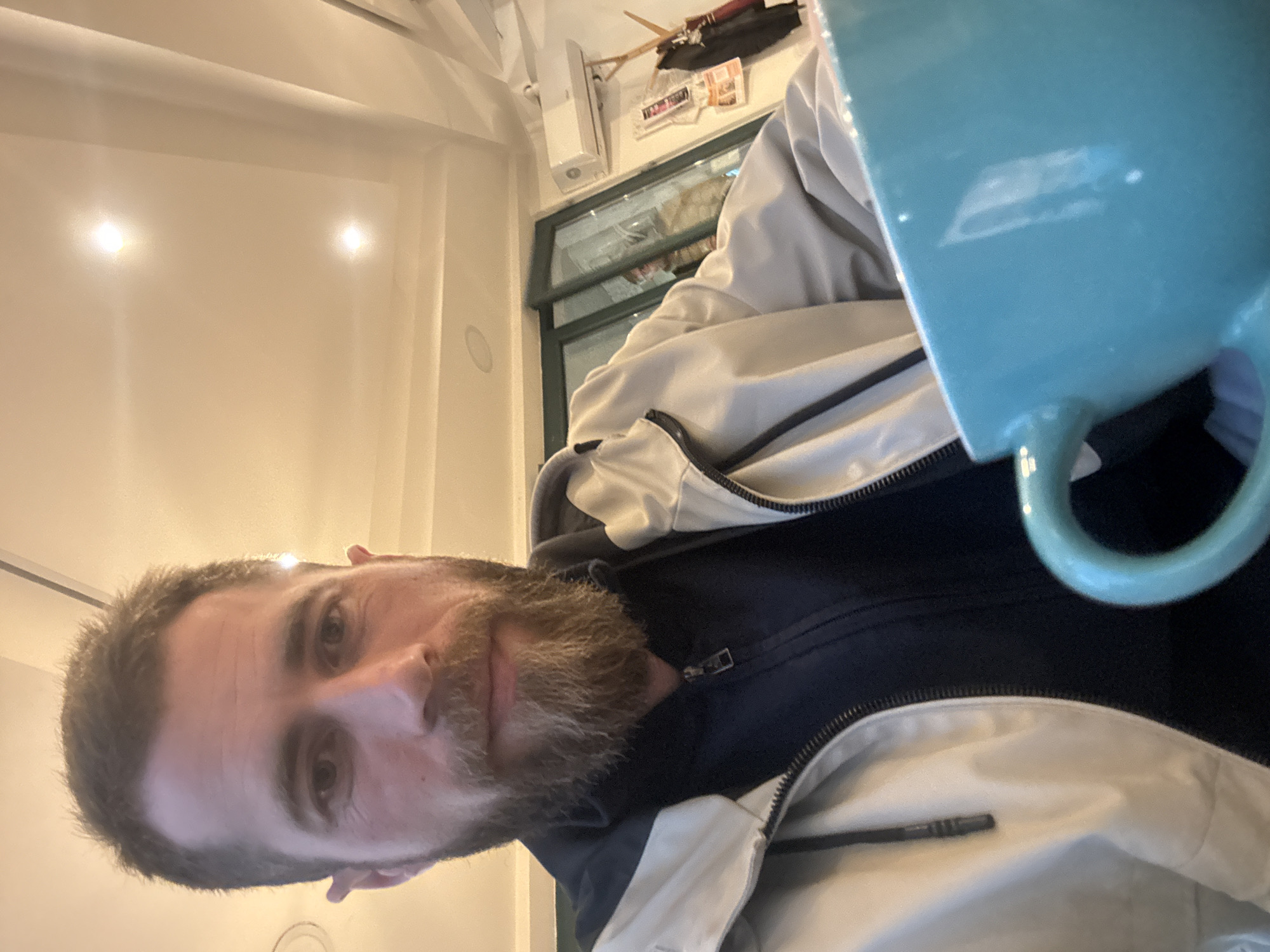 A male individual with short dark hair and a full brown beard is depicted in an indoor setting, looking directly at the camera. He wears a light-colored jacket over a dark, zippered top. A large turquoise blue mug is positioned in the lower right foreground, partially obscuring the subject's body. The background features a light-colored ceiling with recessed lighting, a glass door or window frame, a white wall-mounted air conditioning unit, and a wooden coat rack with a dark garment. Promotional materials are affixed to the wall below the air conditioning unit. The scene is within an indoor commercial establishment, likely a cafe or restaurant. The specific location within Budva, Montenegro, is not discernible from the visual evidence provided.