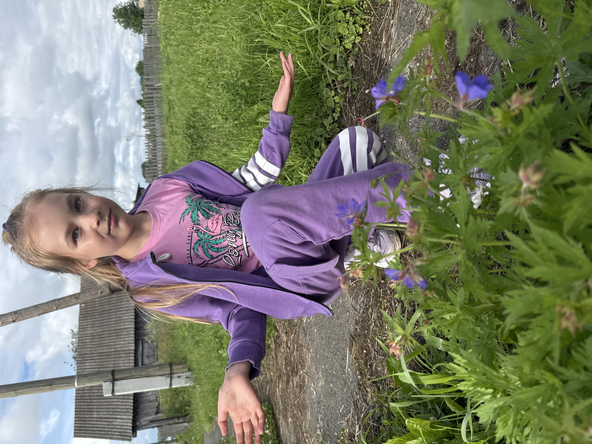 A young girl with blonde hair, wearing a purple hoodie and pants, and a pink t-shirt with a palm tree graphic, is sitting on the ground in an outdoor setting. She is looking towards the camera with a neutral expression. Her right arm is raised, palm facing outwards, as if waving or posing. She is surrounded by lush green grass and wildflowers. In the background, there is a wooden fence and a rustic building, suggesting a rural or village environment. The sky is overcast with clouds. The overall mood is relaxed and casual, possibly during a daytime outdoor activity. Text on the t-shirt is partially visible and appears to say "SEA SURF". The location is identified as Большой Турыш, Russia.