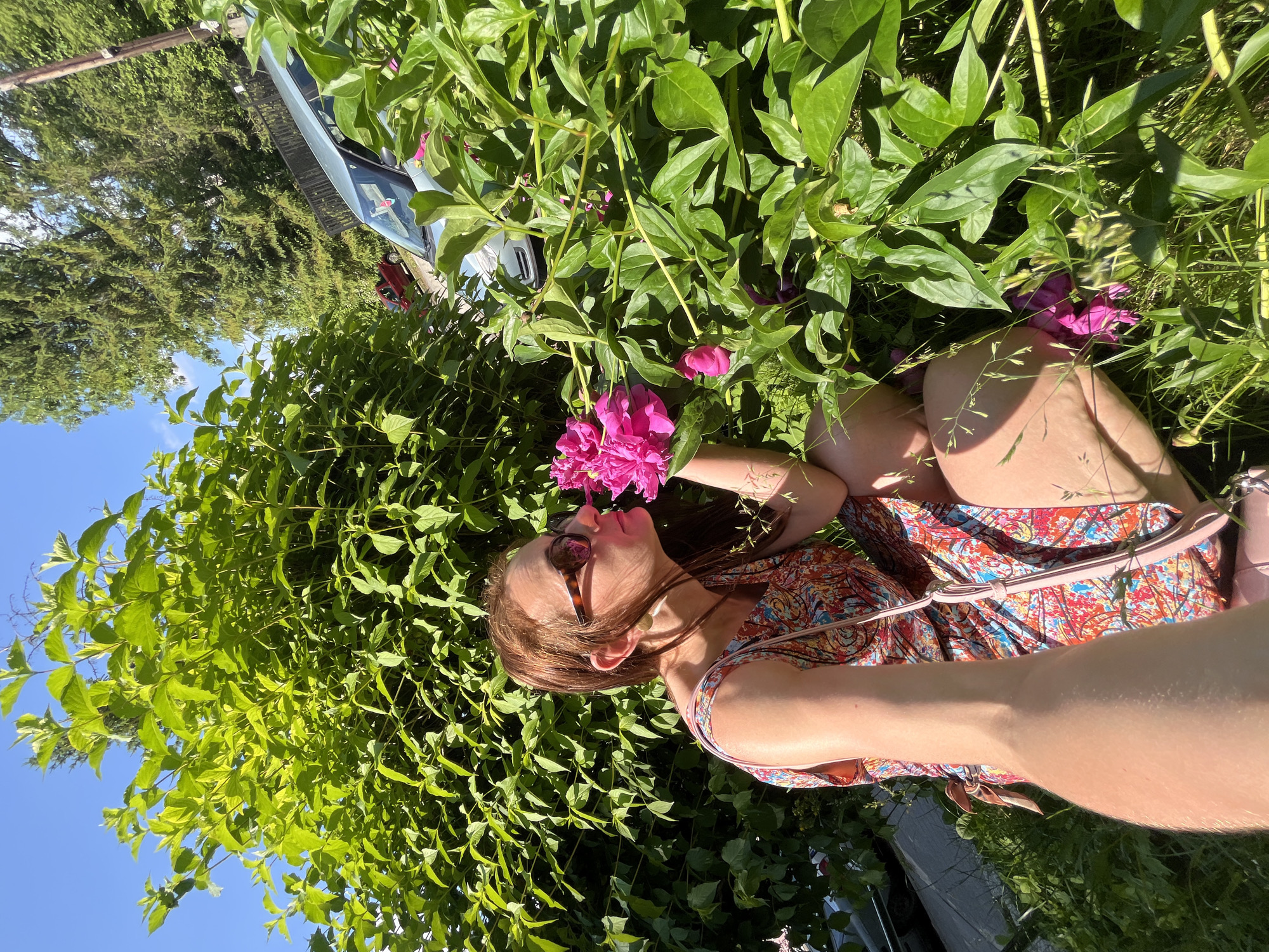 A woman with sunglasses and a colorful dress is kneeling in a garden, surrounded by lush green foliage and pink peonies. She is leaning forward, smelling one of the flowers. Sunlight illuminates the scene, suggesting it is daytime. In the background, a car is parked. The overall mood is serene and peaceful, with the woman appearing relaxed and enjoying nature. The image is a selfie, with the camera angle capturing her from the chest up and her arm extended, holding the camera.