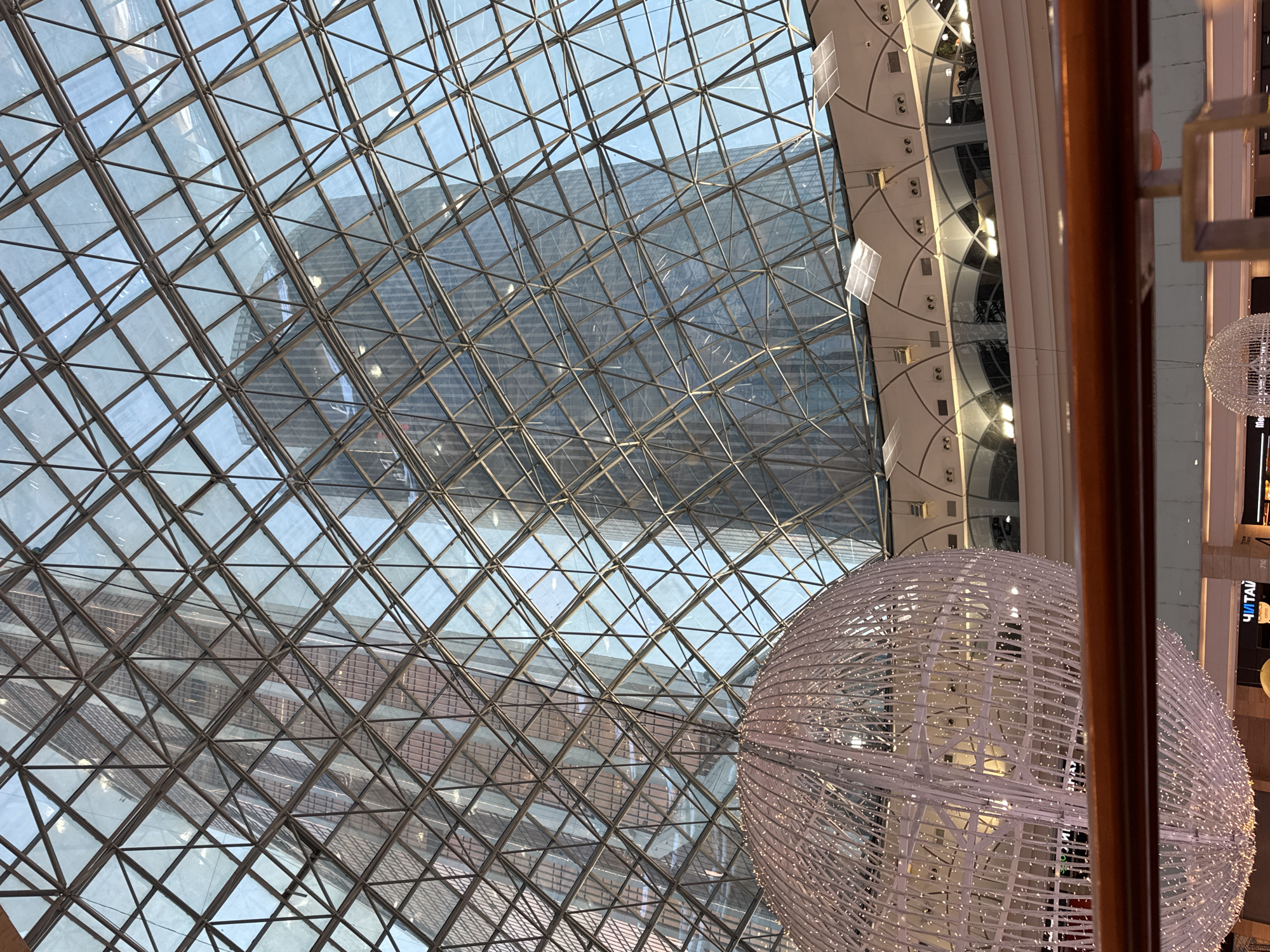The image captures an upward view inside a large, modern building, likely a shopping mall or atrium, in Khimki, Russia. The most prominent feature is the elaborate glass ceiling, constructed from a geometric pattern of metal beams and numerous triangular and diamond-shaped glass panels. Through this transparent roof, a clear blue sky is visible, along with reflections of a modern skyscraper or large building, showing its structured facade with rows of windows.
To the right, a massive spherical light fixture, made of a white, intricate wireframe structure adorned with countless small lights, hangs from the ceiling. This sphere occupies a significant portion of the lower right frame. Above and behind it, a curved section of the interior ceiling, possibly part of an upper floor or balcony, is visible. It features recessed lighting and several square, translucent panels. A wooden or wood-toned railing runs vertically along the right edge of the frame, indicating the photo was taken from an elevated position.
The bright lighting suggests it is daytime with clear weather. On a dark background behind the spherical light, partially obscured by the railing, white Cyrillic text is visible, clearly showing "ЧИТАЙ" (Chitay, meaning "READ"). Below this, another part of a logo or text, possibly "DLC", can be discerned. The overall impression is one of modern architecture, spaciousness, and bright, inviting interiors.