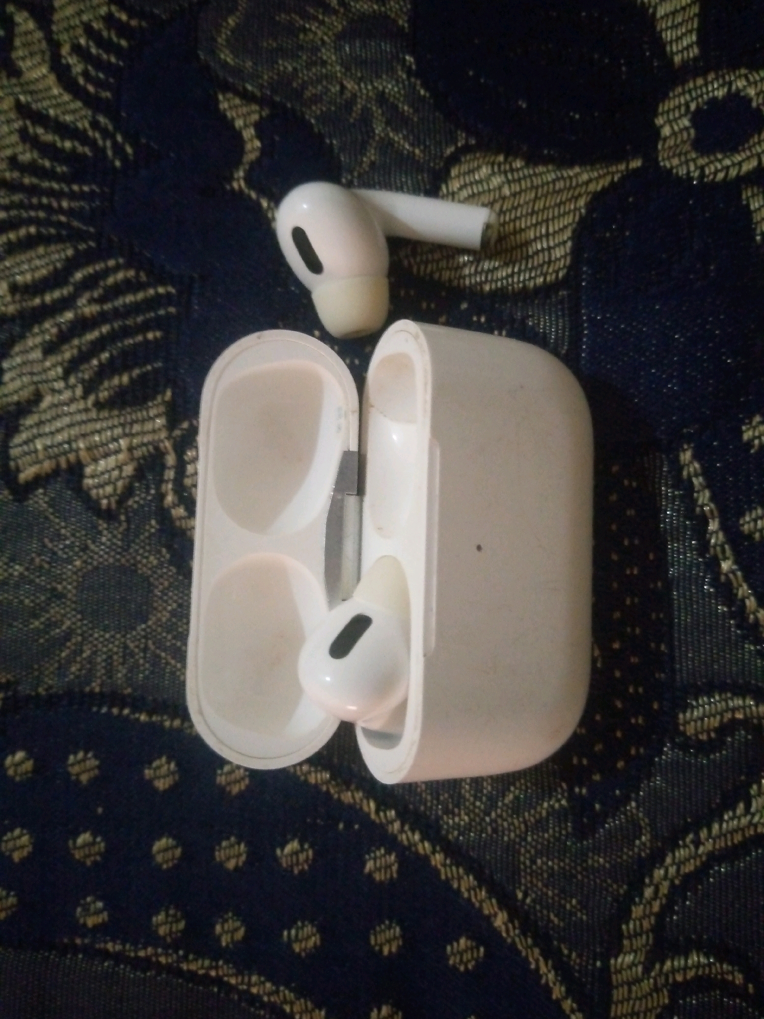 The image displays a pair of white wireless earbuds and their charging case resting on a dark blue fabric with a woven pattern of gold and black floral motifs. One earbud is placed on top of the open charging case, while the other earbud is partially visible, extending to the left of the case. The charging case is open, revealing two indentations where the earbuds are meant to sit. The lighting in the image appears to be artificial and somewhat dim, casting subtle shadows. The location is identified as Khalilpur, Pakistan.