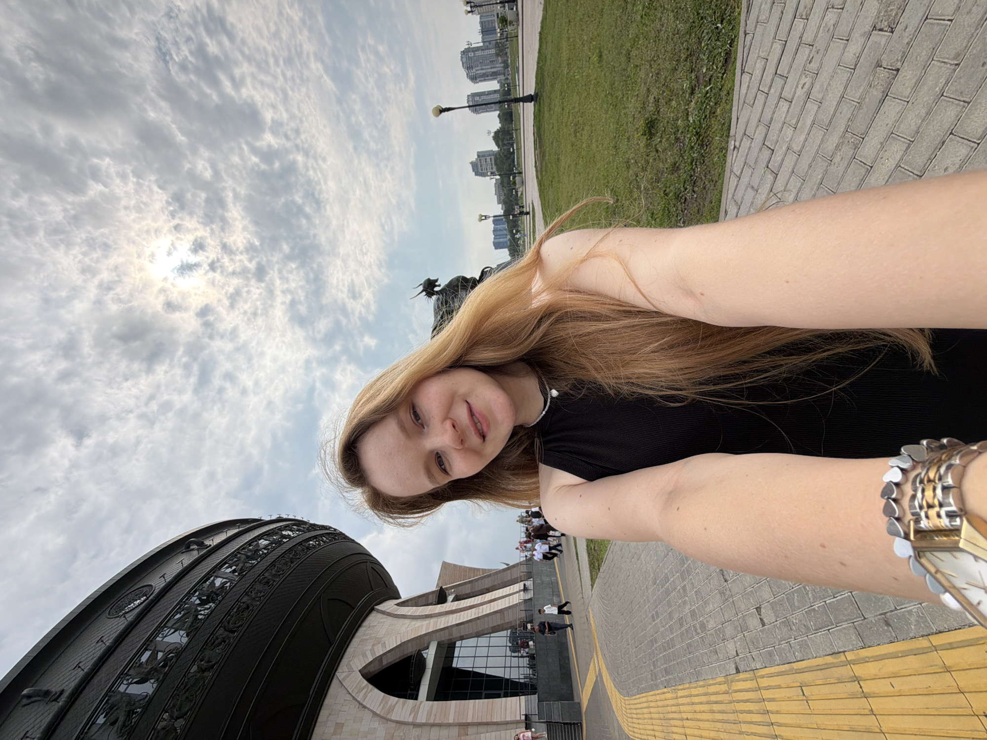 A young woman with long, blonde hair is taking a selfie outdoors. She is wearing a black tank top and a silver necklace. She is standing on a paved walkway with a yellow tactile paving strip to her left. In the background, to her left, is a large, circular building with a dark, ornate facade. The building has a wide base with arched entrances and a tiered upper section with decorative railings. To the right of the building, there is a grassy area with trees and lampposts. In the distance, there are several tall, modern buildings. The sky is partly cloudy with the sun shining through. A statue of a dragon-like creature is visible behind the woman on the right side of the frame. The scene appears to be in a public urban space. The woman's right arm is extended, holding the camera, and she is wearing a watch with a metal band on her left wrist. Several other people are visible walking in the background, near the entrance of the building. This location is identified as the "Temple of All Religions" in Kazan, Russia.