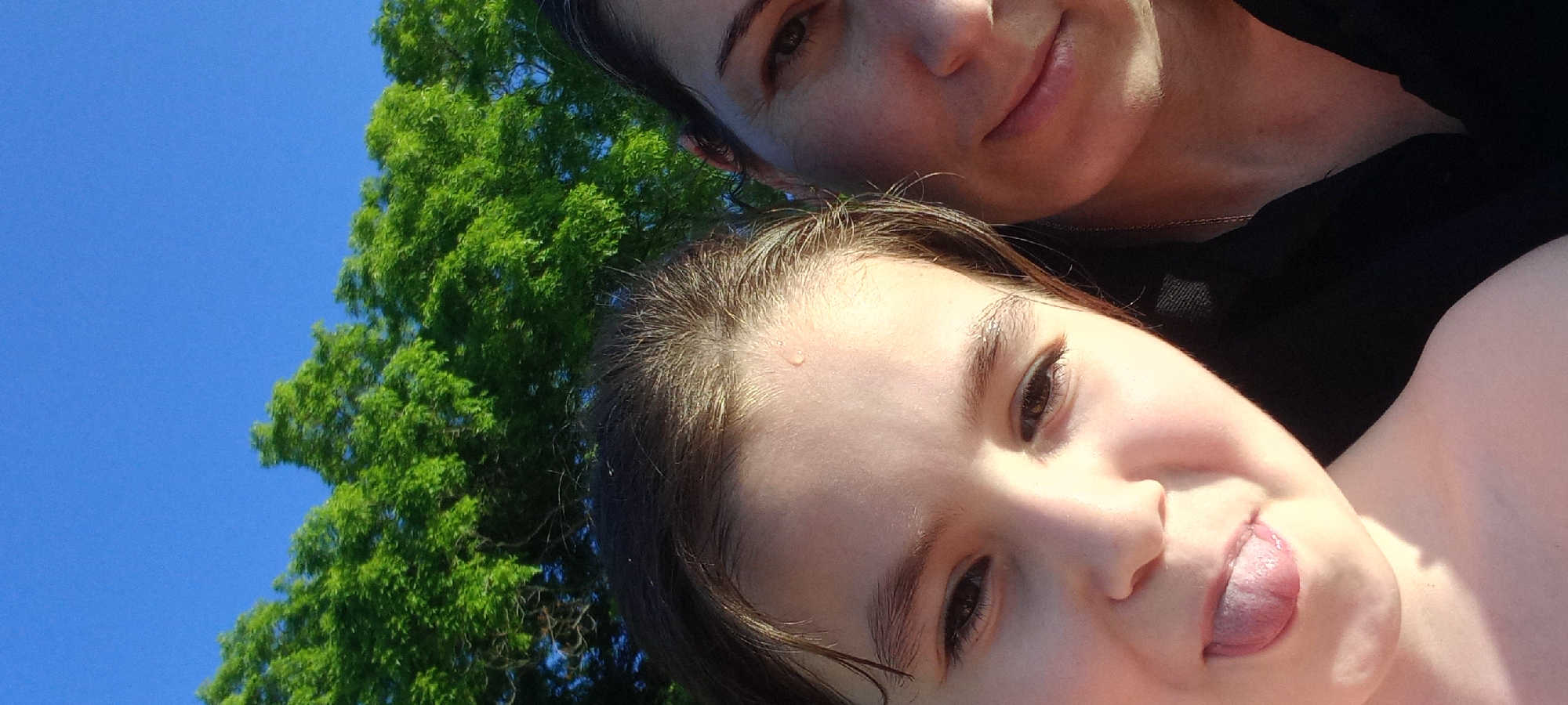 This is a close-up, selfie-style photograph of two people with bright green trees and a blue sky in the background. The person on the left, a younger individual, has dark hair and is sticking out their tongue, with a small droplet of water visible on their forehead. The person on the right, an adult, has dark hair and is smiling, with a delicate gold chain visible on their neck. Both individuals appear to be outdoors. The location of Gwandra, Abkhazia is not visible or inferable from the image.