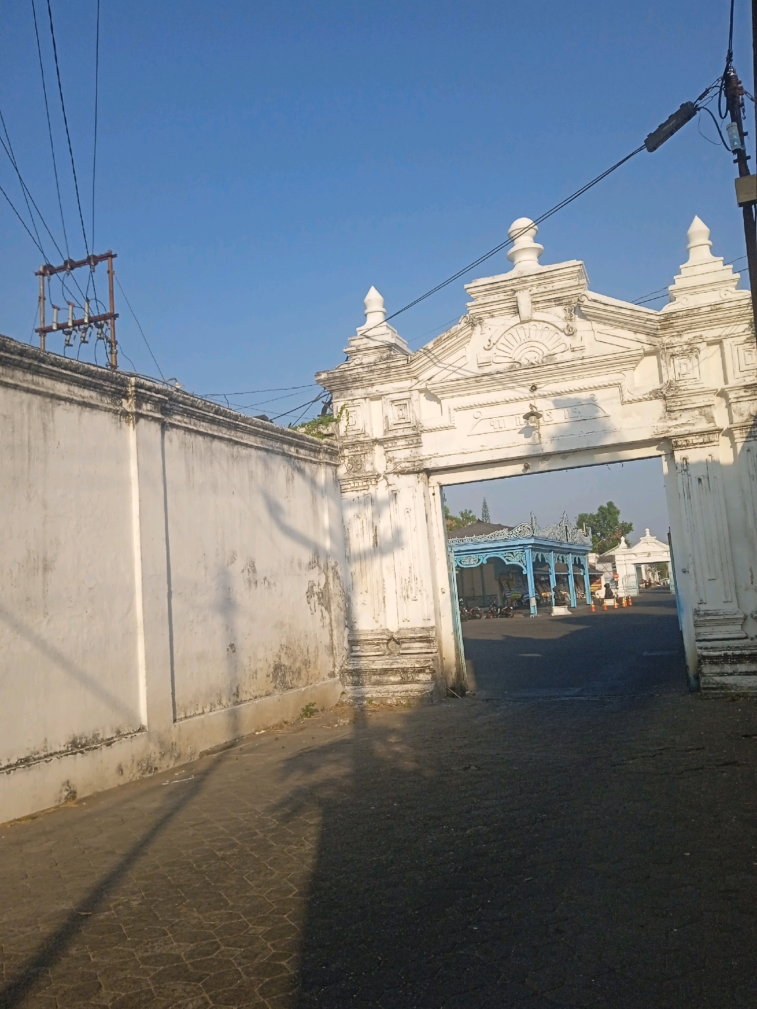 An ornate, white arched gateway stands in the foreground, leading into a courtyard. The gateway is decorated with classical architectural elements, including columns, carved friezes, and finials. To the left of the gateway is a long, white, weathered wall, a portion of which appears to be constructed from stone. A transformer pole with electrical wires is visible above the wall and the gateway.
Through the archway, a blue pavilion with intricate carvings is visible in the courtyard. The courtyard itself is paved and appears to be a public space. Several motorcycles are parked in the courtyard, and a few people can be seen in the distance. Orange traffic cones are also present in the courtyard. The sky is a clear, bright blue. The paving stones in the foreground are hexagonal. The scene is bathed in the warm light of what appears to be early morning or late afternoon sun, casting long shadows across the ground.
This gateway is located at the entrance to the Surakarta Sunan's Palace complex in Surakarta, Indonesia.