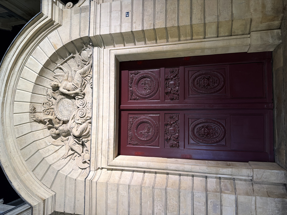 The image displays the grand entryway of the Hôtel de Boisgelin, which houses the Polish Embassy, located at 47, rue de Longchamp, in the 16th arrondissement of Paris, France.
The scene presents a large, ornate double door set within a light-colored, classical stone facade, illuminated by artificial light against a dark background. Above the doors, an arched stone pediment features a detailed central relief sculpture. This sculpture depicts two draped, winged female figures, commonly identified as allegorical figures or angels, positioned symmetrically on either side of a central, empty circular escutcheon. The figure on the left holds an upward-reaching leafy branch, while the figure on the right holds a long, downward-pointed straight horn or trumpet. A small carved wreath with ribbons is situated above the escutcheon.
The double doors are painted a deep burgundy or dark red. Each door panel features three distinct recessed and carved sections. The uppermost sections are circular reliefs; the left depicts a seated female figure, and the right displays another seated figure. The middle sections are rectangular reliefs, characterized by ornate scrollwork and two cherubic figures supporting a central oval shield. The lowermost sections are oval reliefs, each containing a carved grotesque mask or human-like face.
On the stone facade to the immediate right of the door frame, a small black plaque with the white number "47" is affixed. No individuals are visible in the scene.