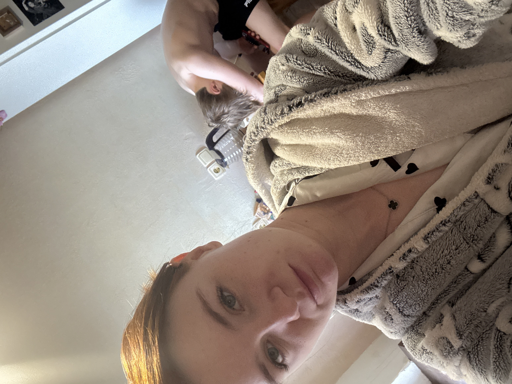 The image shows a person, presumably female, taking a selfie. She is wearing a grey and white patterned bathrobe. A silver necklace with a clover-shaped pendant is visible. In the background, another person, likely a child, is bending over. They are wearing black shorts and have light brown hair. A white electrical outlet and a clear plastic container are also in the background. The environment appears to be a bedroom. There is no information about the city of Sinyavino, Russia.