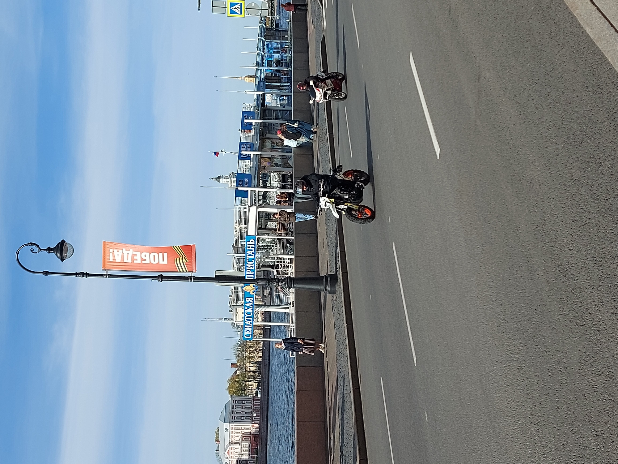 The image shows a street scene in Saint Petersburg, Russia. A two-lane road is on the right, with two motorcycles traveling in the same direction. On the left of the road, there's a pedestrian walkway bordered by a river or canal. People are walking along the walkway. Banners are attached to the street lamps, featuring a message in Russian related to Victory Day ("ПОБЕДА!"). Buildings are visible in the background across the river or canal. A sign reads "СЕНАТСКАЯ ПРИСТАНЬ" in Russian. The sky is blue with some clouds.