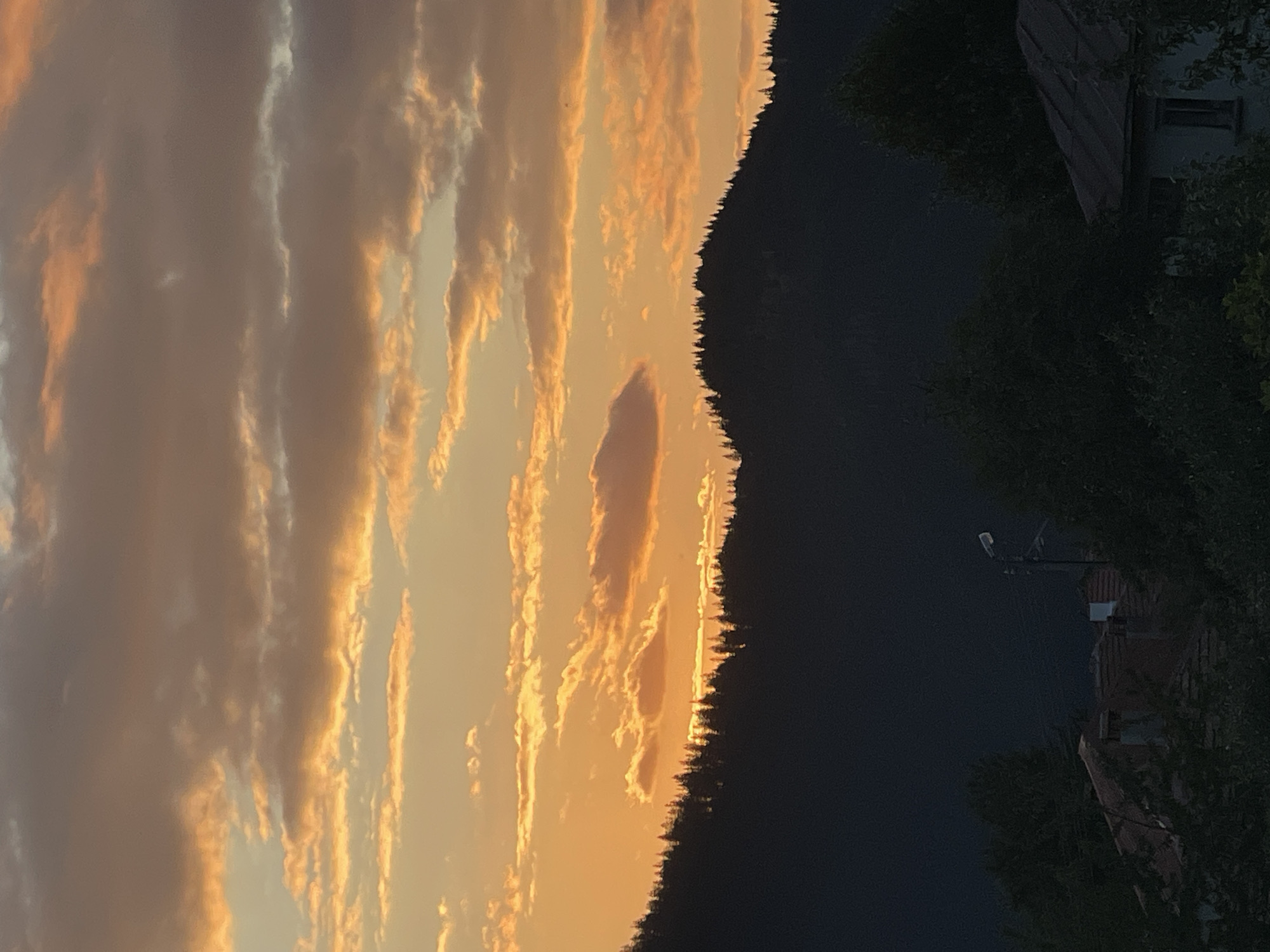 This is a landscape photograph of a sunset over a mountain range near Manastir, Bulgaria. The sky is filled with dramatic clouds illuminated by the setting sun, casting hues of orange and yellow. A dark silhouette of a forested mountain ridge runs horizontally across the middle ground, with the trees forming a jagged outline against the vibrant sky. Below the mountain, in the foreground, are the dark outlines of houses and trees, suggesting a village or settlement nestled at the foot of the mountains. The overall impression is one of a serene and picturesque natural scene at dusk.