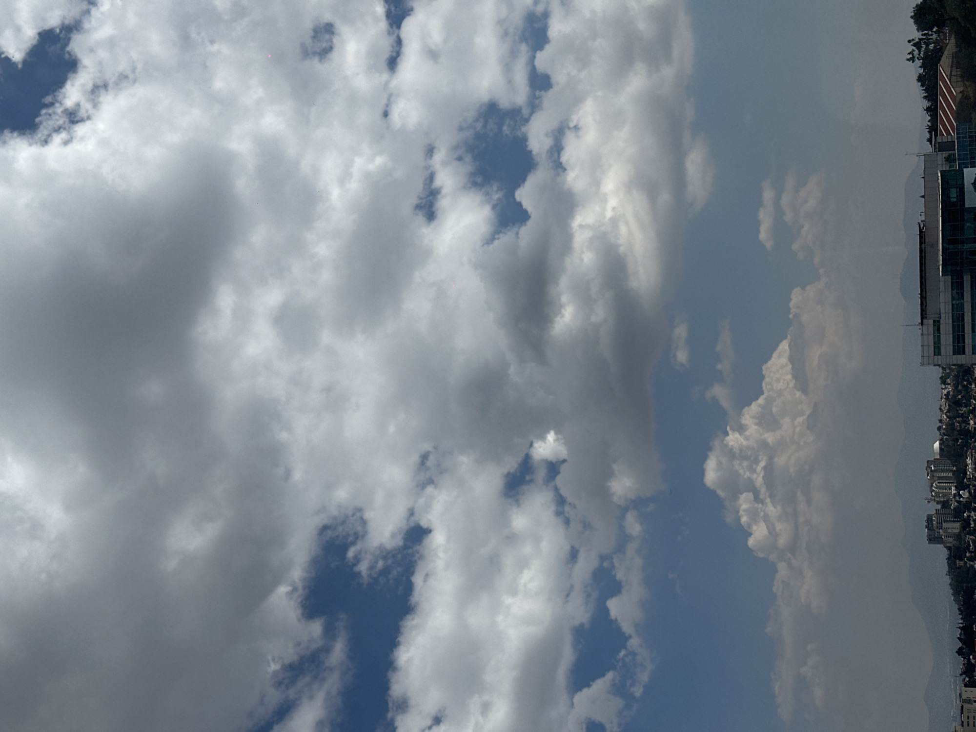 The image depicts a cloudy sky over a part of Huixquilucan de Degollado, Mexico. The sky is filled with cumulus clouds of varying shades of white and gray, with patches of blue sky visible in between. On the right side of the image, a cityscape is visible, with buildings and structures extending along the terrain. A cluster of buildings and an American flag are visible at the upper right. The terrain appears to be a mix of residential and possibly commercial areas.
