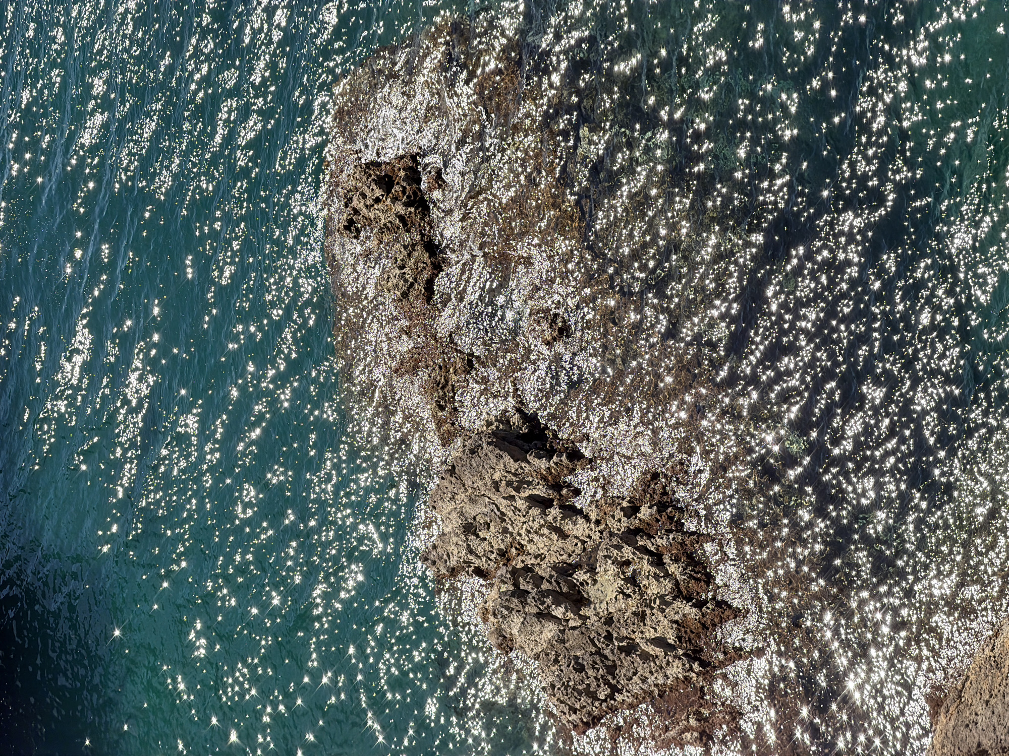 A high-angle shot shows a rocky outcrop extending into the sea at Mirište, Montenegro. The water is teal and speckled with sunlight reflections. The rocks are porous and partially submerged.